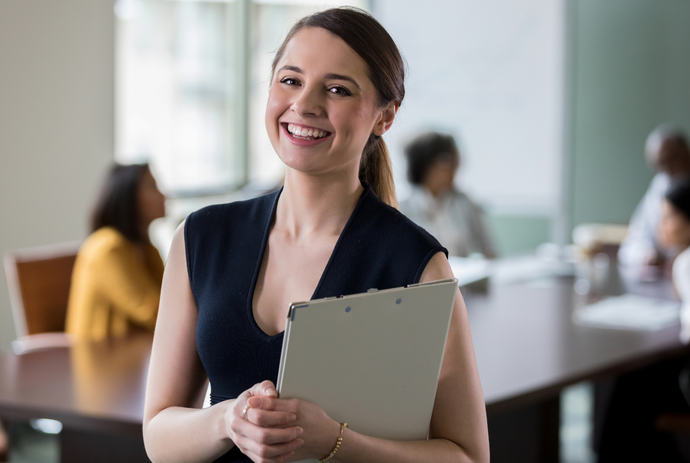 A woman is smiling while holding a clipboard in an office.