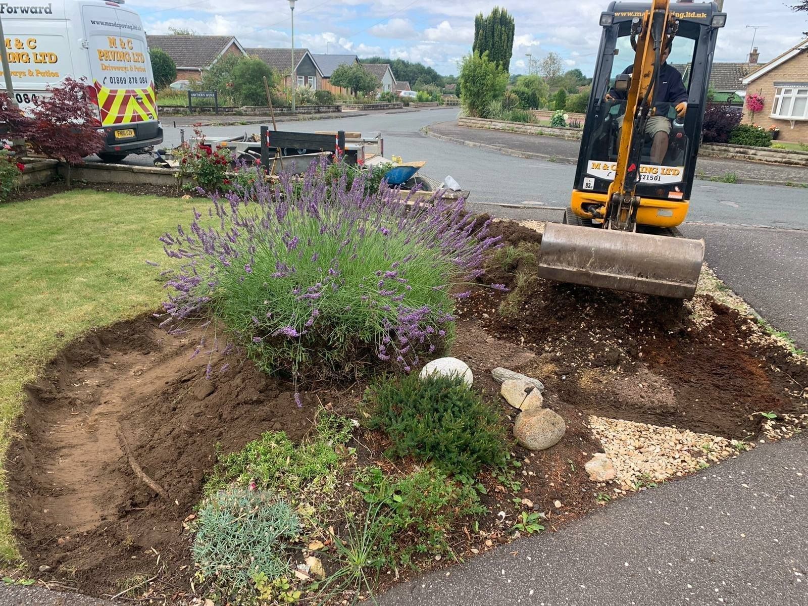 Excavator clearing soil around a landscaped flowerbed beside a road and parked truck