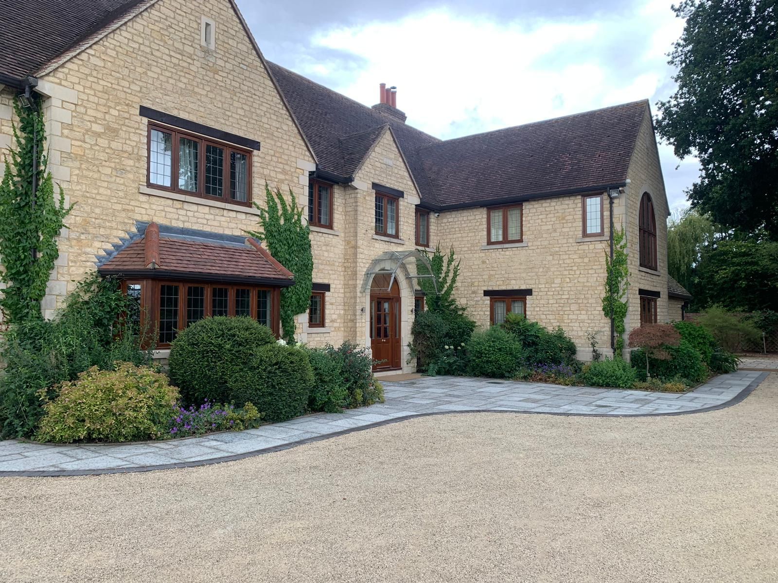 Large beige stone house with brown roof, arched doorway, and gravel driveway on a quiet street