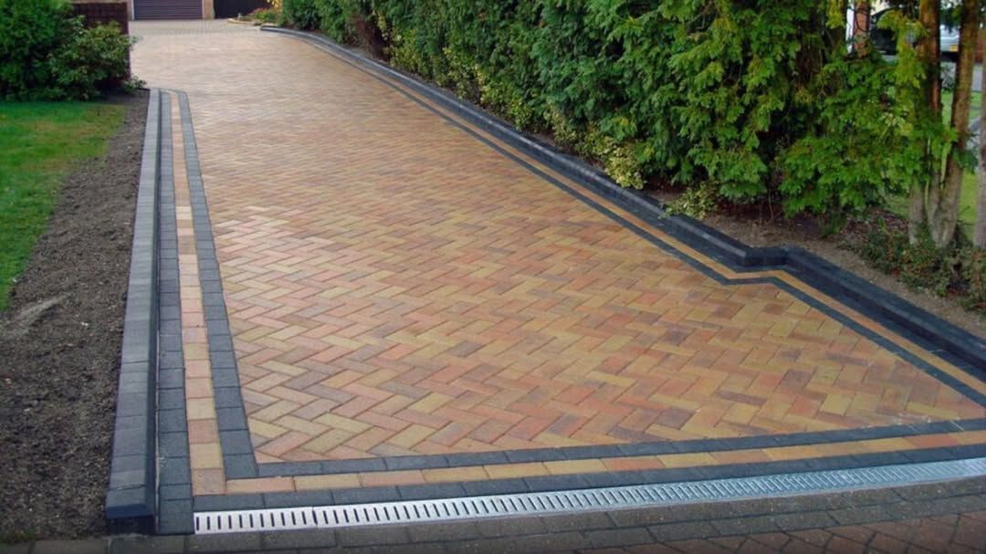 Brick-paved driveway bordered by dark edging and hedges, with a drainage grate at the front