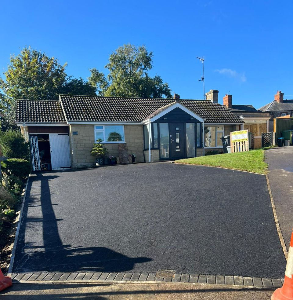Modern single-story house with a large black driveway, white walls, and blue sky in front.