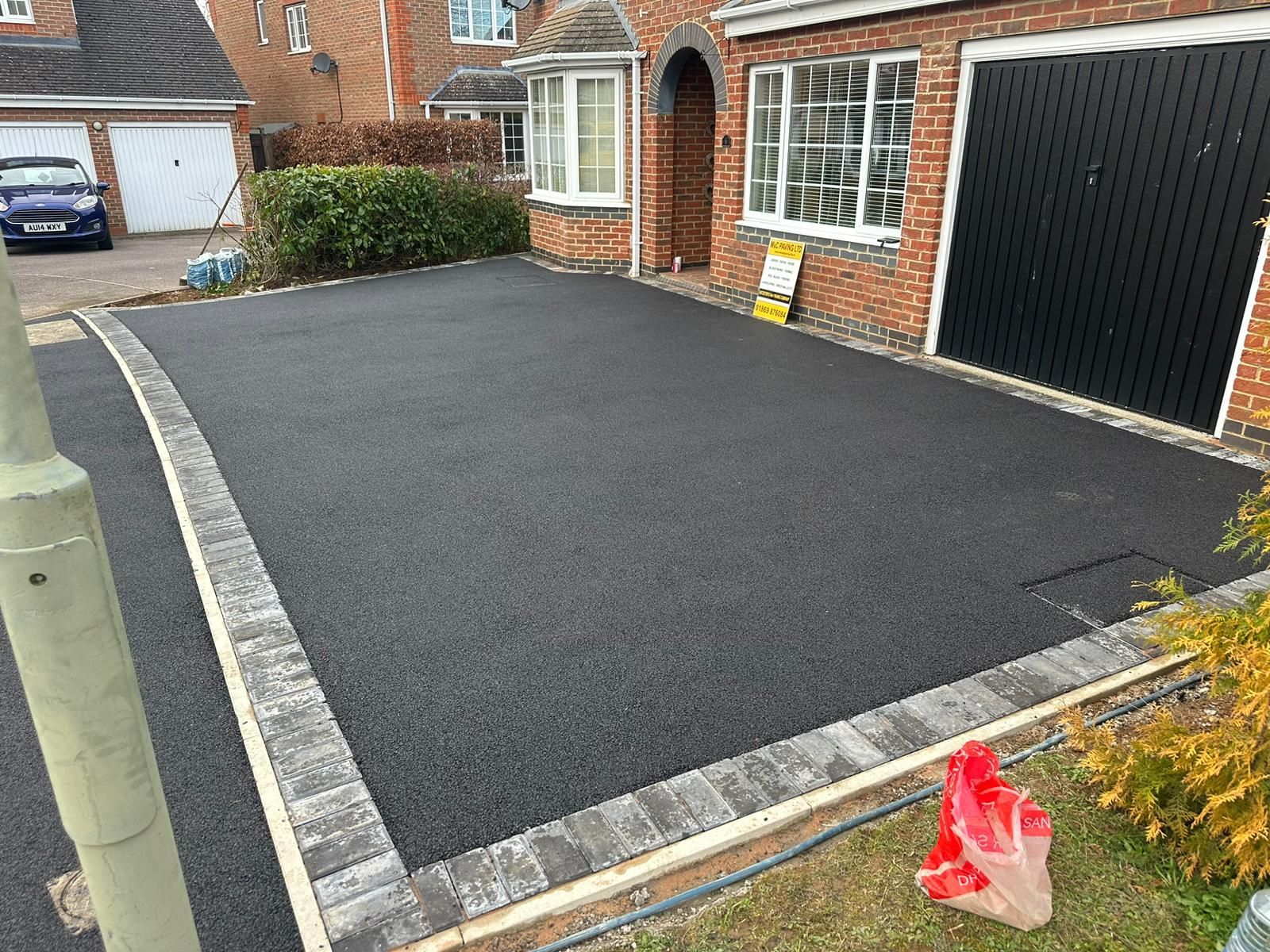 Freshly paved black driveway beside a brick house and garage, edged with light gray pavers.