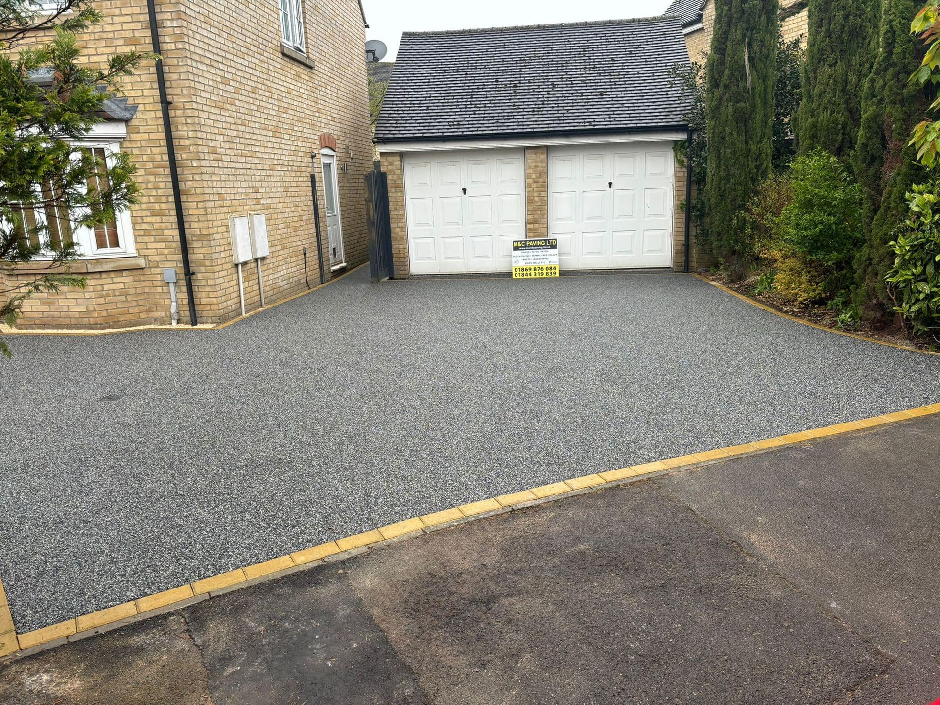 Gravel driveway leading to a white double garage beside a stone house and greenery