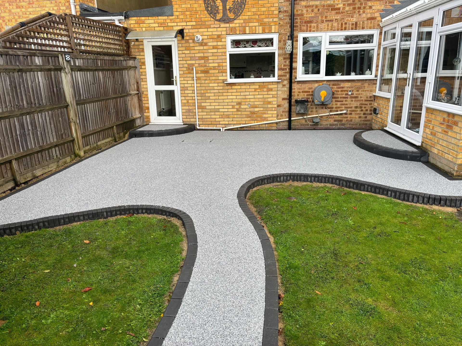 Backyard patio with gray gravel paths, green grass, and a brick house with windows and a door.
