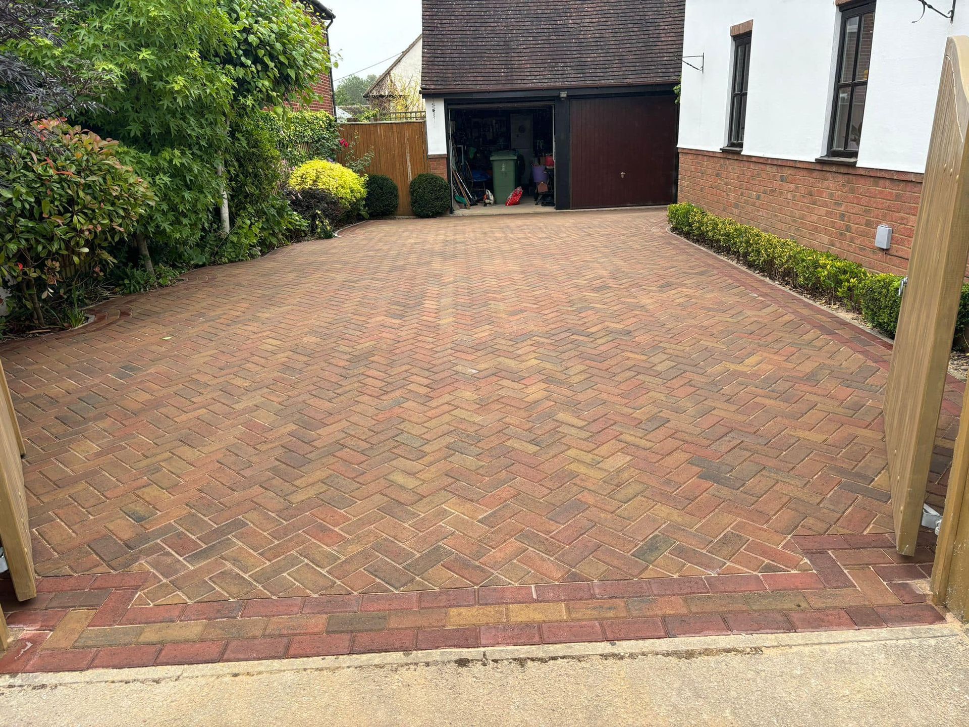 Brick-paved driveway leading to a dark garage, bordered by hedges and a brick wall