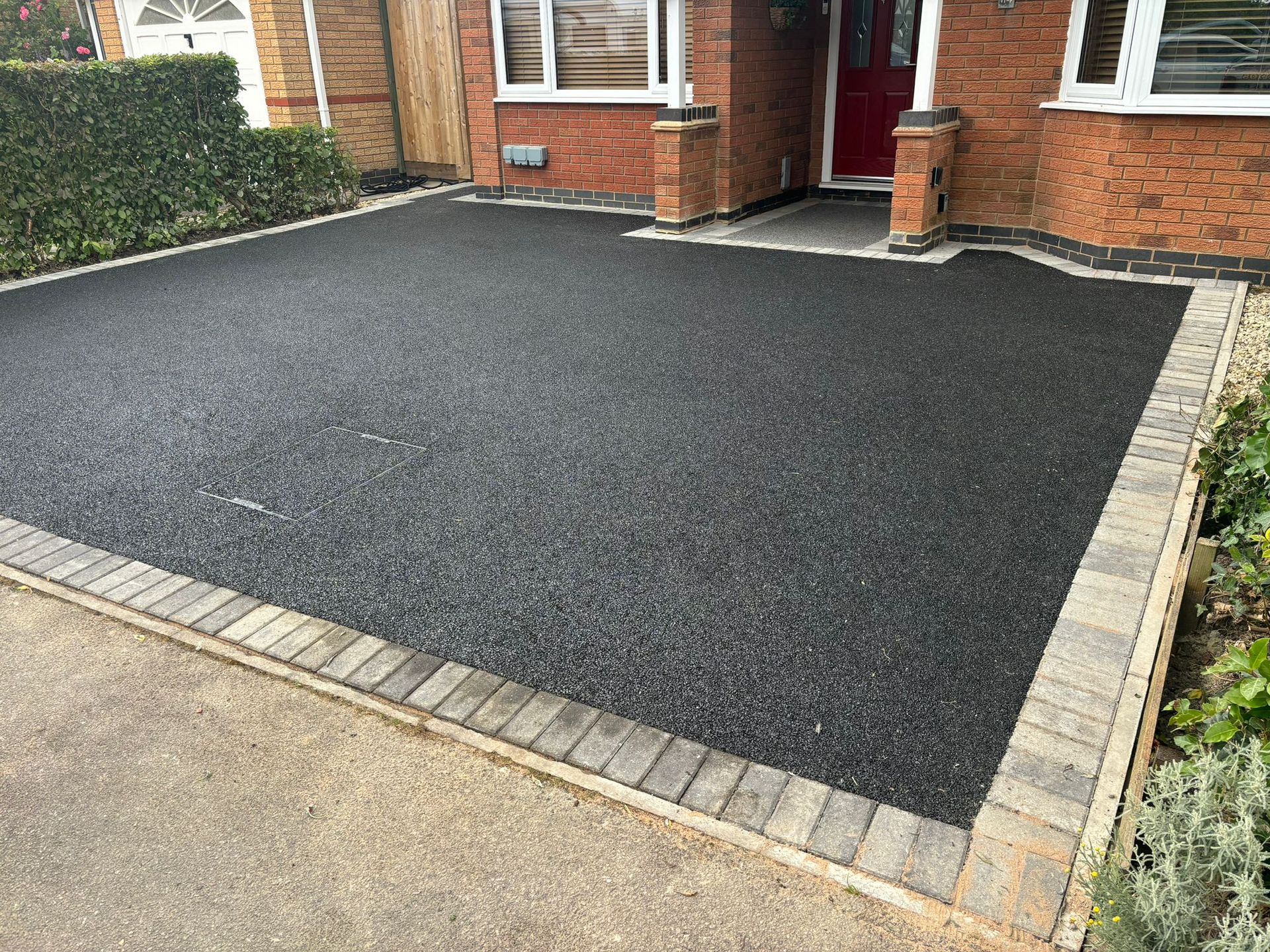 Freshly paved black driveway in front of a brick house with light stone edging, viewed from the street