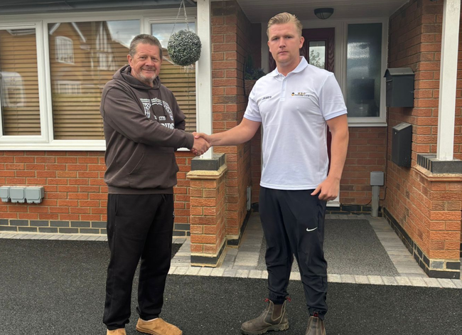 Two men shaking hands outside a brick house, one holding a key near the front door