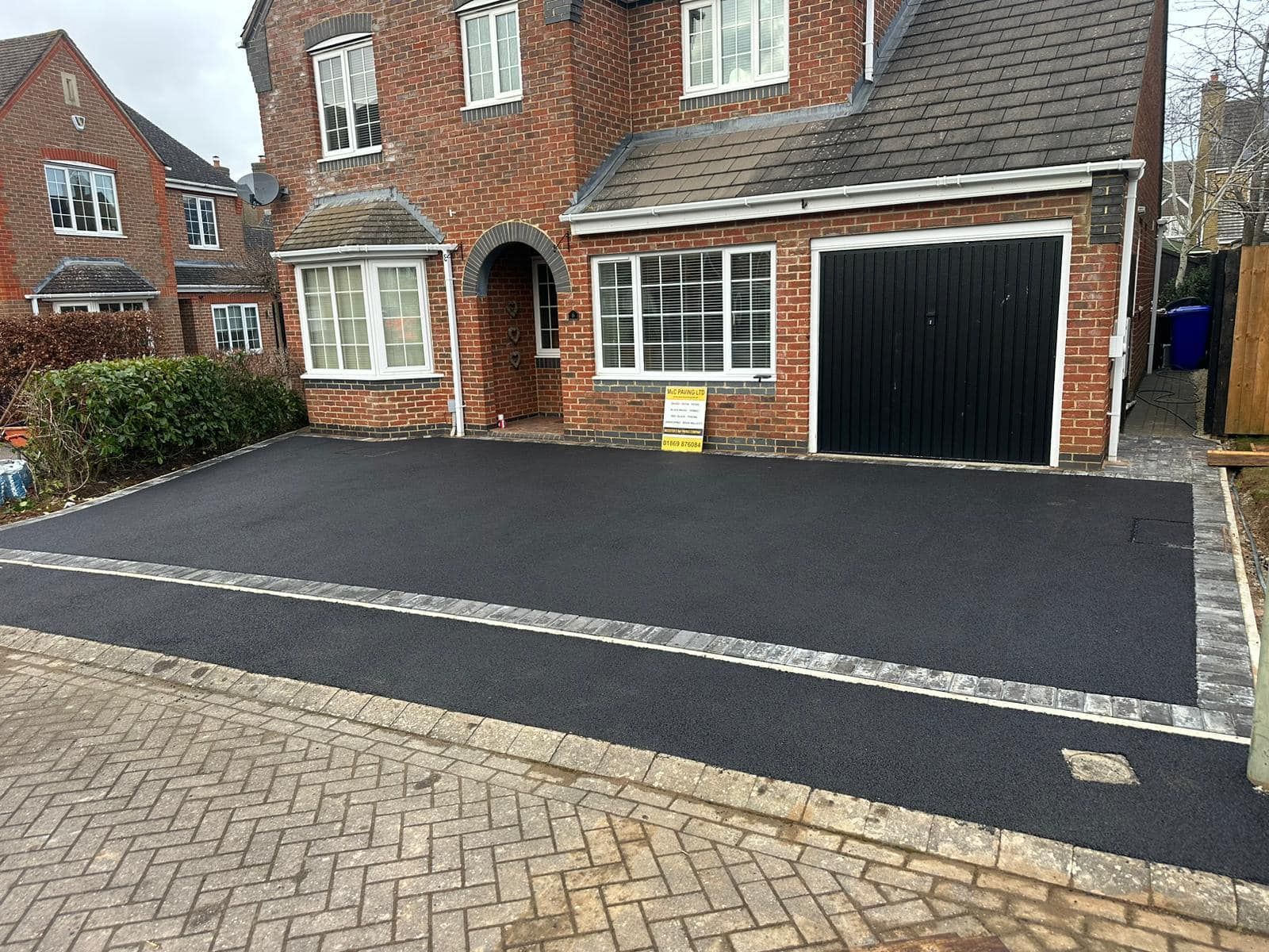 Freshly resurfaced black driveway beside a brick house with a garage, viewed from the street.