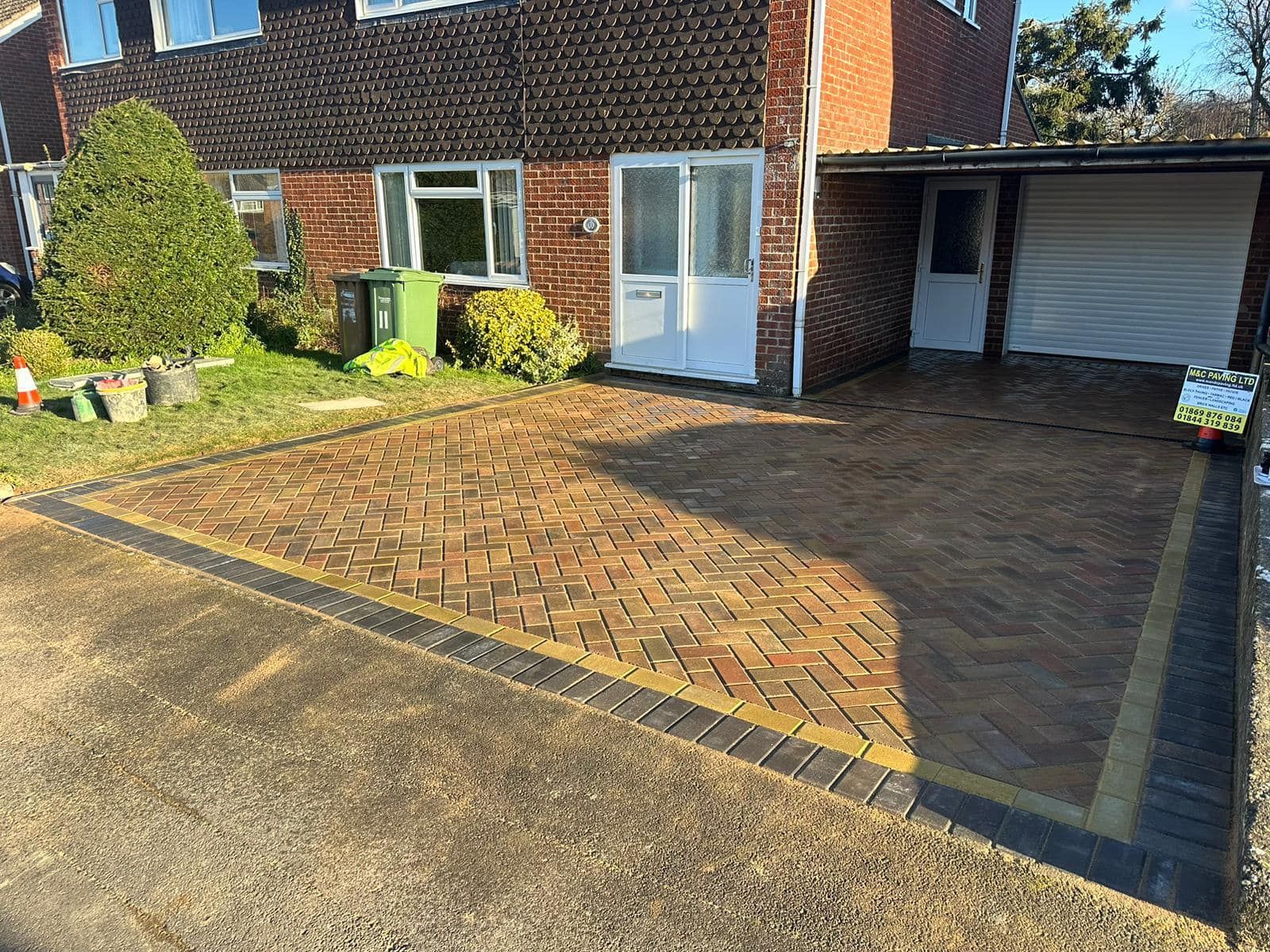 Brick house front with a paved driveway and white patio doors, bordered by a small garden and hedge.
