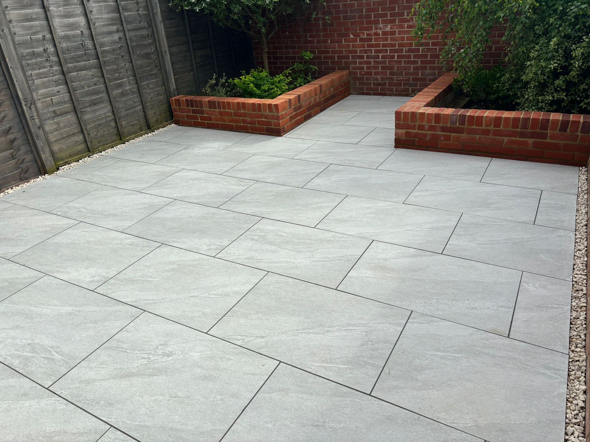Empty paved patio with light gray tiles, brick planters, and a garden wall.