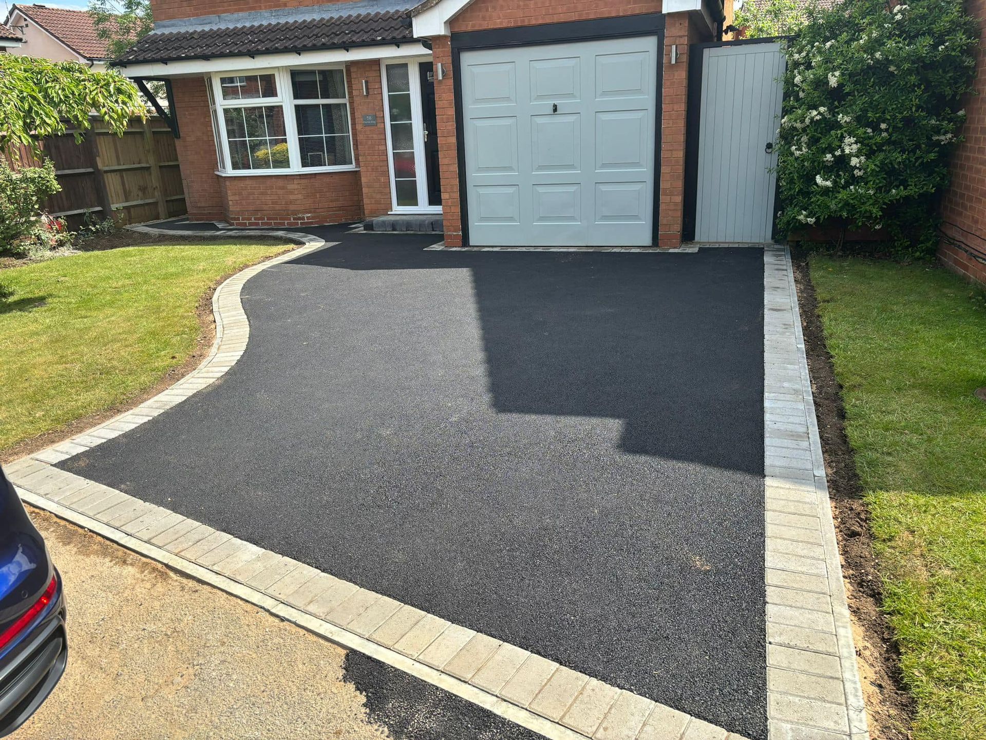 Freshly paved dark driveway leading to a garage beside a brick house with lawn and hedges