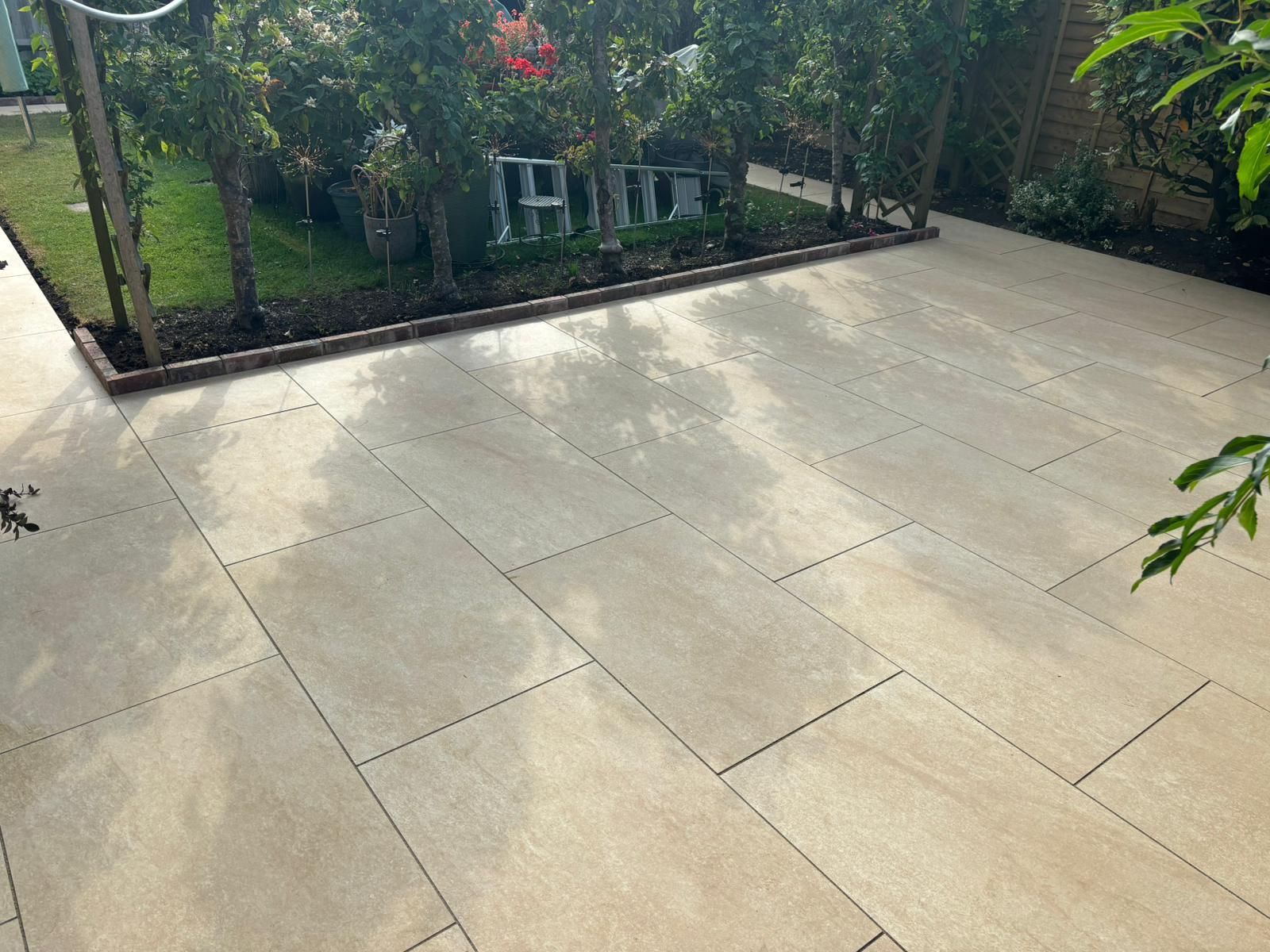 Large beige-tiled patio bordered by garden greenery and trees, with a house in the background