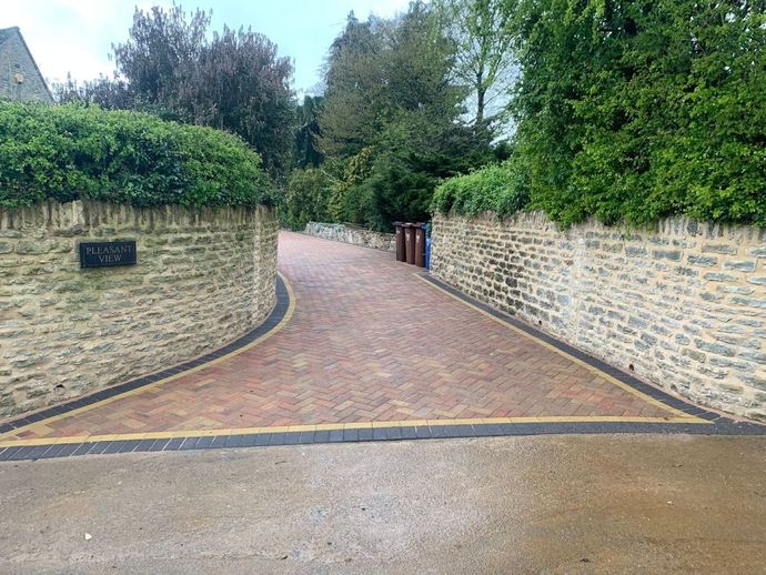 Brick driveway between stone walls, curving toward a gate with trees overhead.