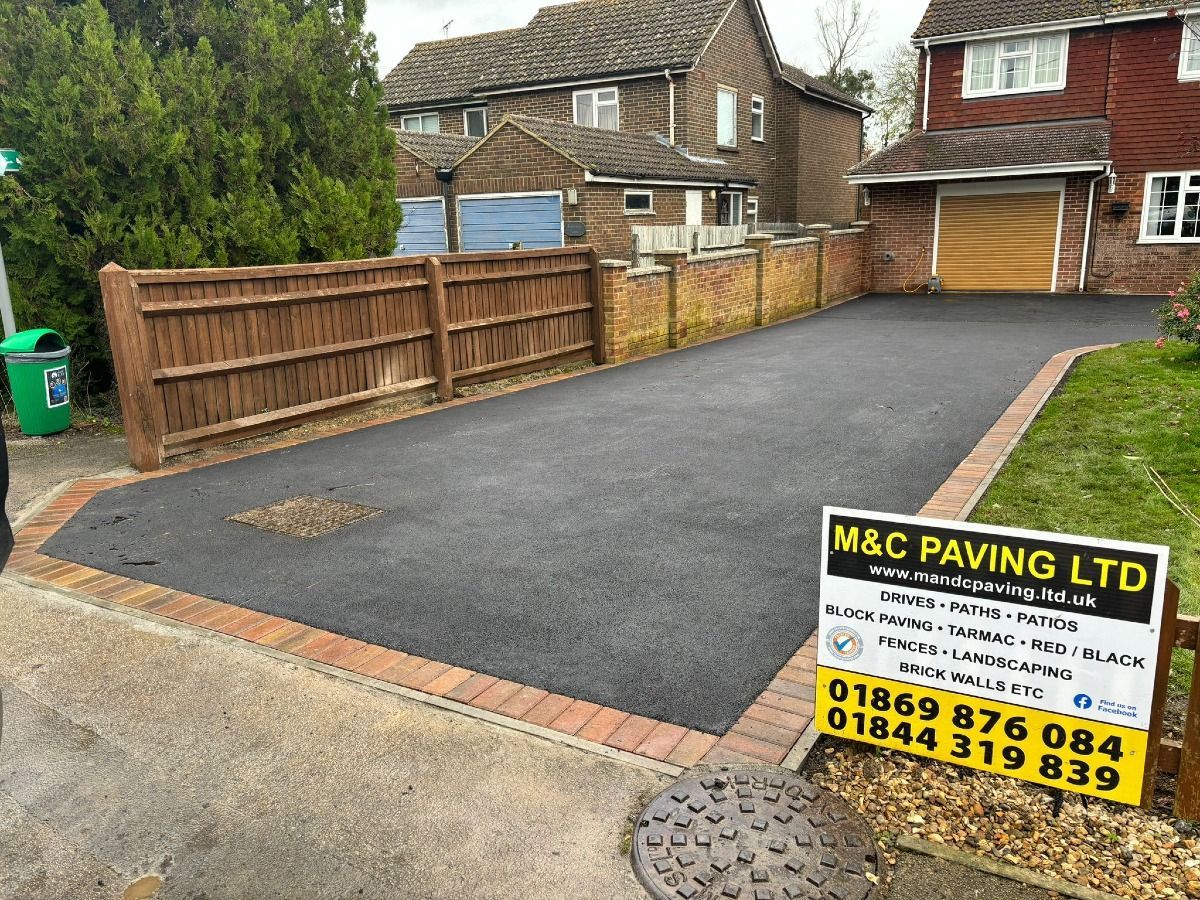 Freshly paved driveway beside wooden fences and houses, with an M&C Paving Ltd sign in the foreground