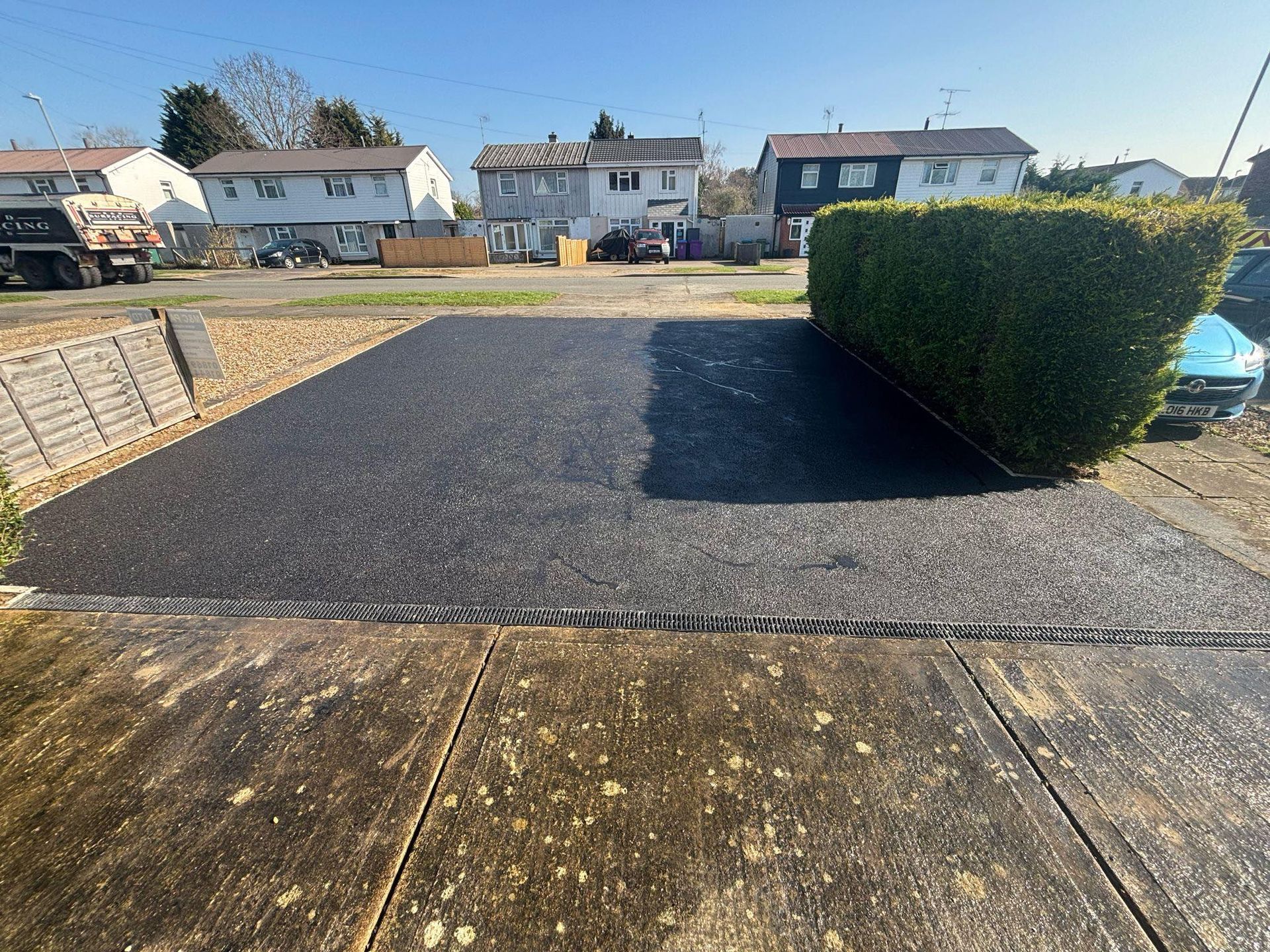 Suburban driveway and front yard with a hedge, gravel path, and houses in the background.
