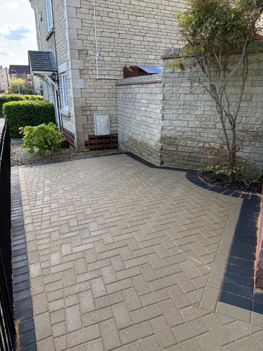 Brick-paved walkway beside a stone house and shrubs, with a small bench and curved dark edging.