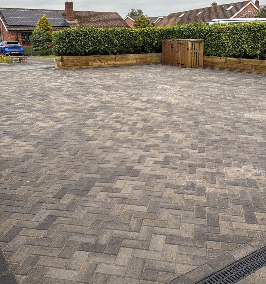 Empty paved driveway with herringbone brick paving, low hedges, and wooden fencing under overcast sky
