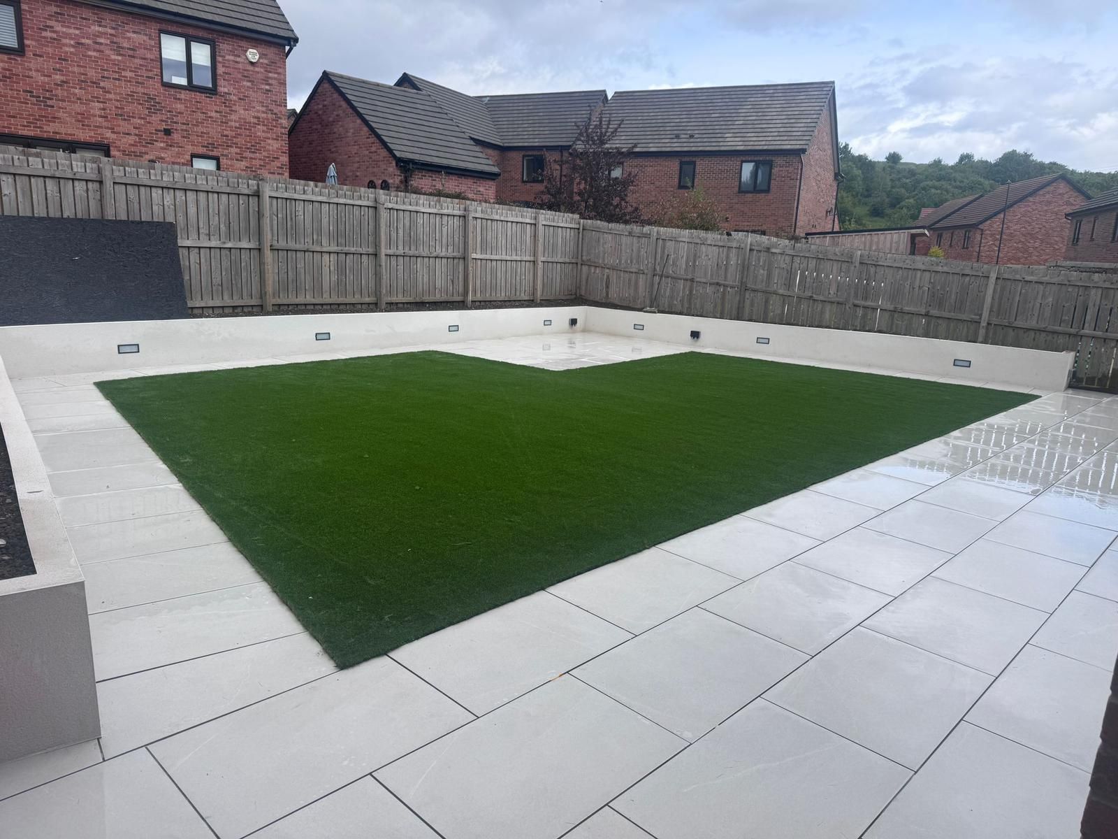 Backyard patio with white paving and a central green artificial lawn, bordered by wooden fencing and brick houses