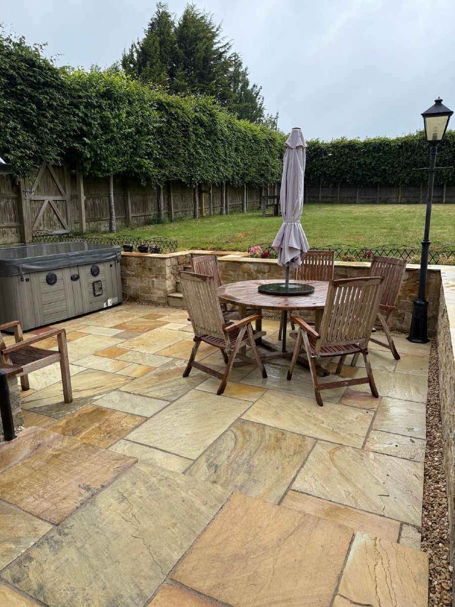 Patio with stone paving, outdoor table and chairs, closed umbrella, grill, and hedge-lined lawn