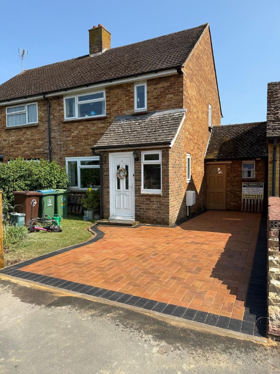 Brick semi-detached house with orange driveway, white front door, and side gate under a sunny sky