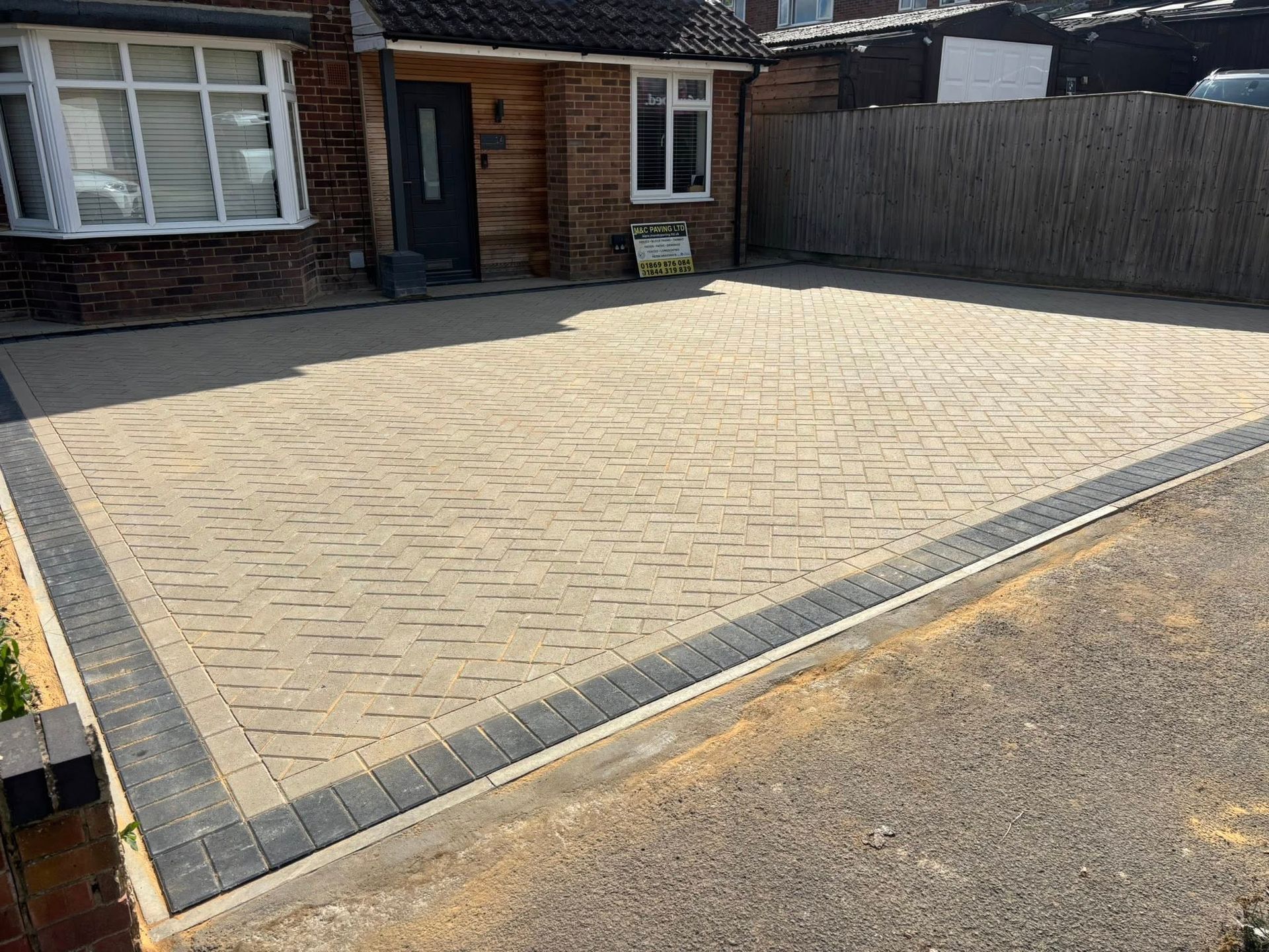 Paved driveway with light brick pavers and dark border in front of a house with a wooden fence