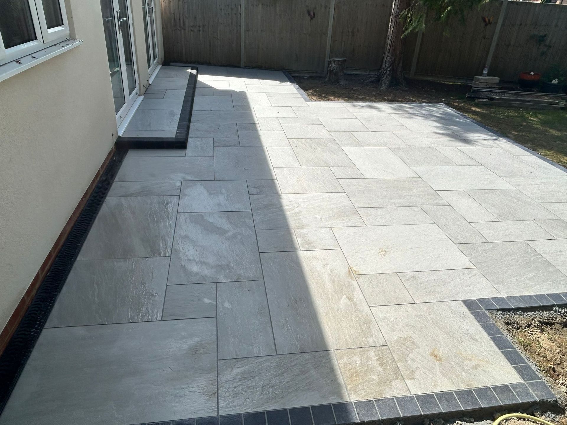 Sunlit stone patio walkway beside a house and fence, with a shaded corner and garden bed edge