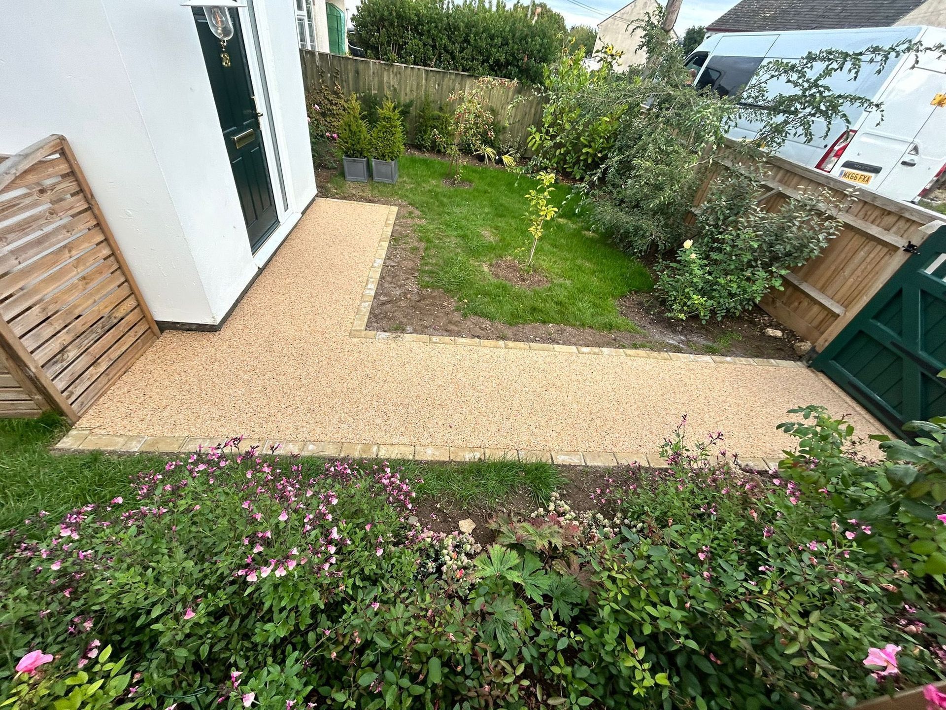 Small backyard with gravel path, green lawn, shrubs, and a white house beside a wooden fence.