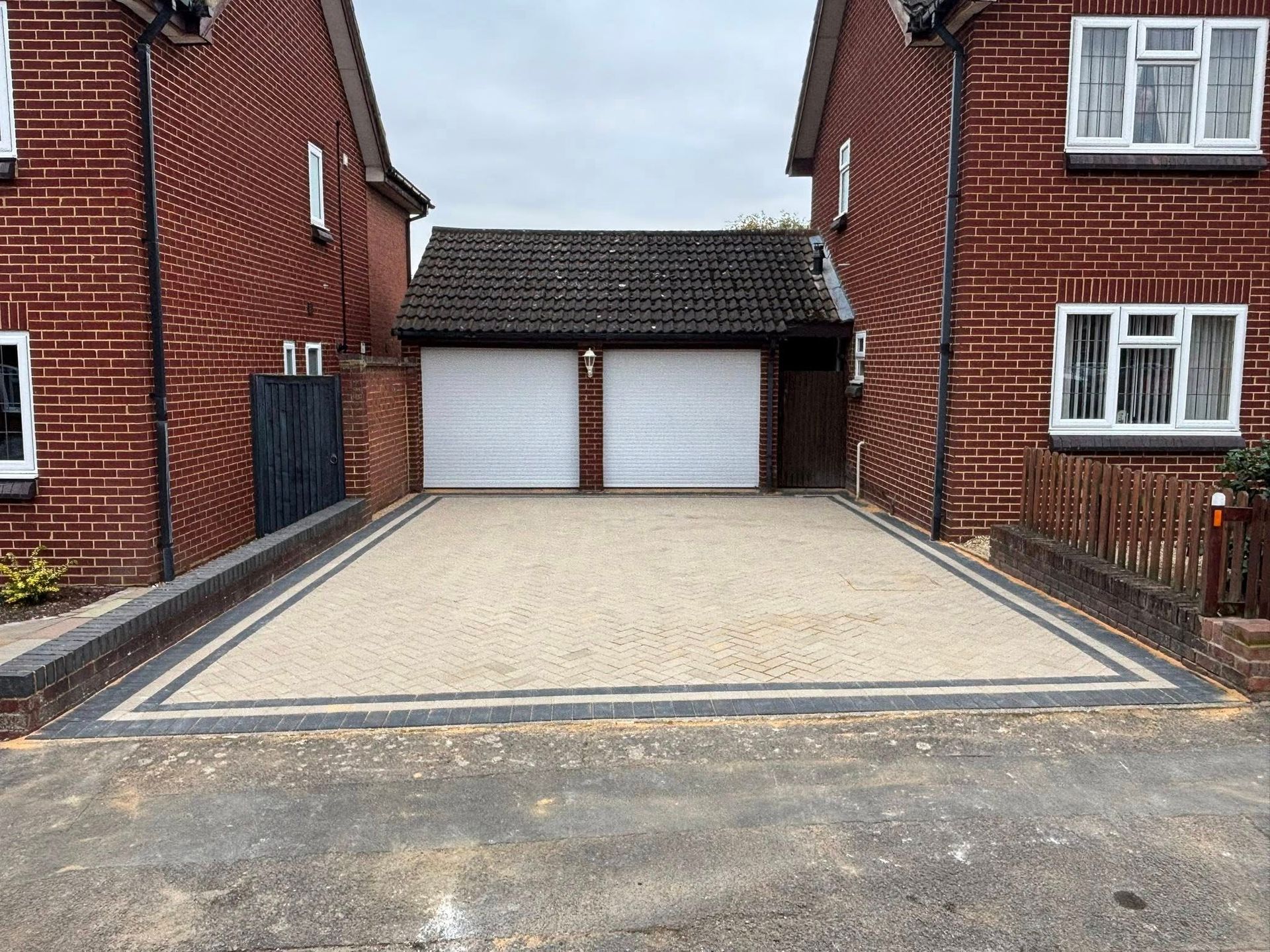 Driveway between brick houses leading to a double garage with white doors and a gravel surface.