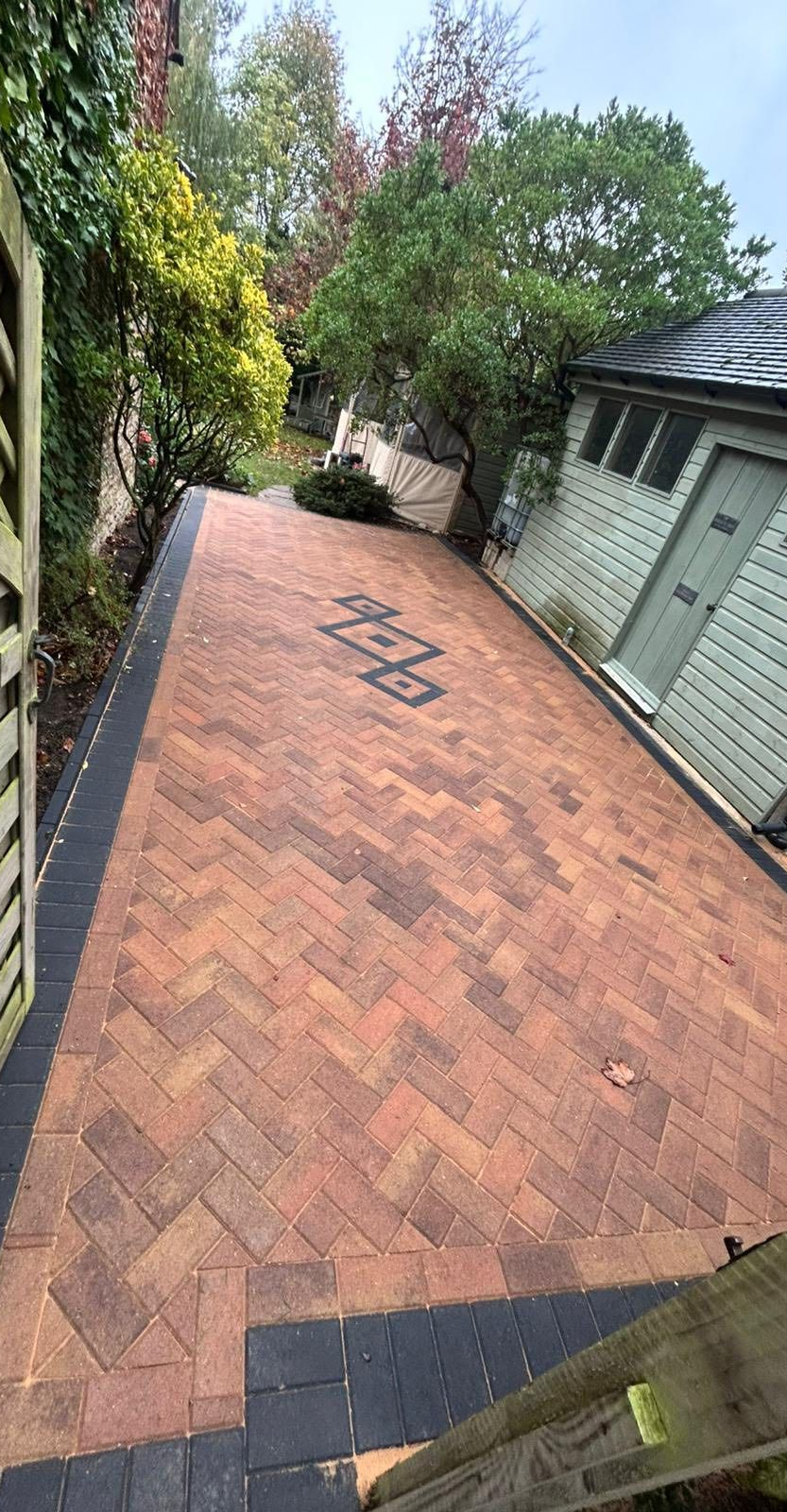 Brick driveway with a tiled house wall and green trees lining the side, viewed at an angle