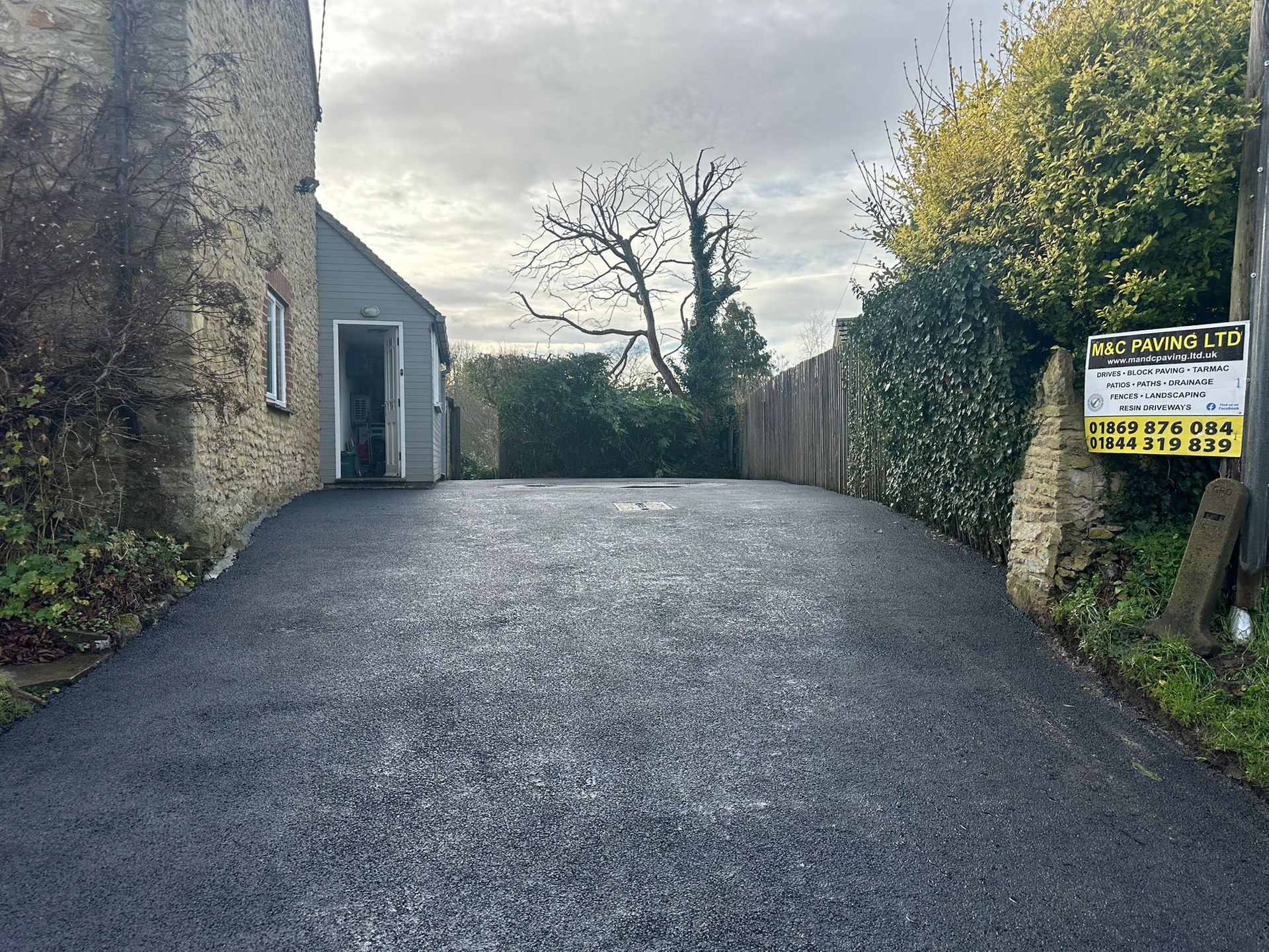 Steep paved driveway beside stone building and hedges, with a gate and sign at the top.
