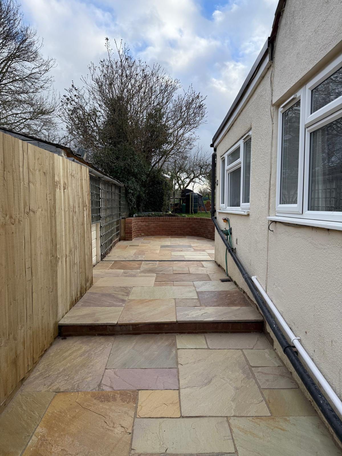 Stone patio walkway beside beige house and wooden fence leading to a raised flower bed under a cloudy sky