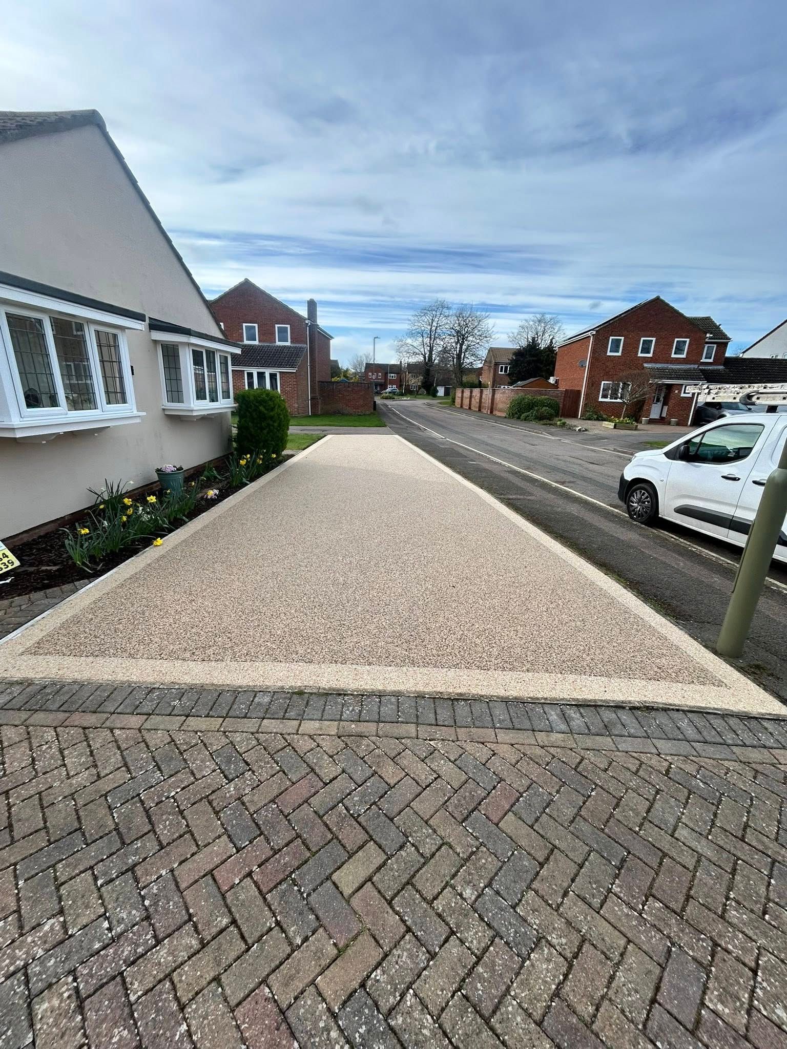 Gravel driveway beside a white house on a suburban street with parked cars and brick homes.
