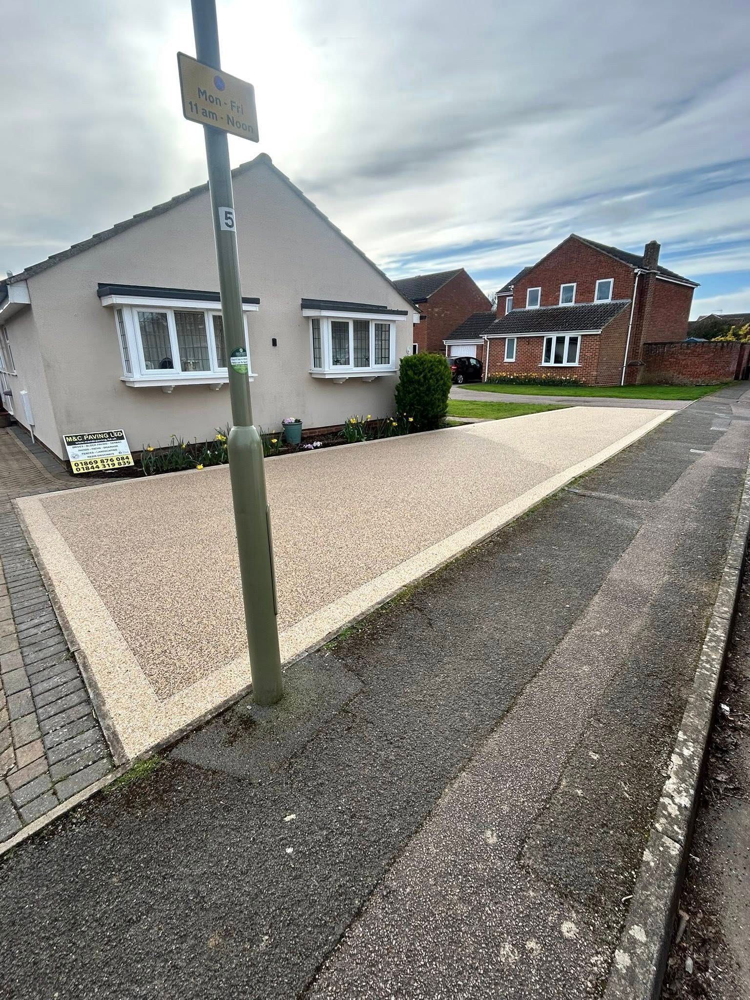 Quiet residential street with detached houses, gravel driveways, and a signpost under a cloudy sky