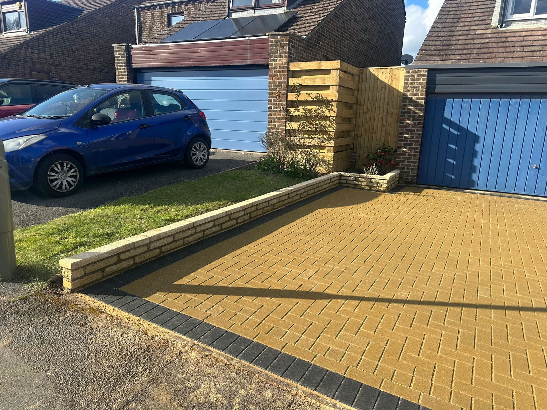 Blue car parked beside a brick-paved driveway and garage with a low wall under construction.