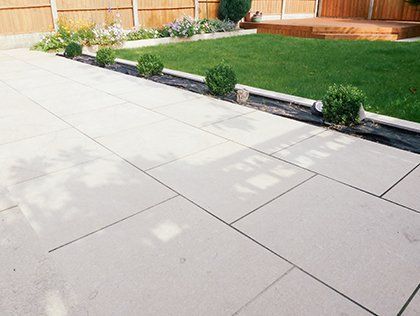 Backyard patio with large light-gray pavers, small shrubs, green lawn, and a wooden fence