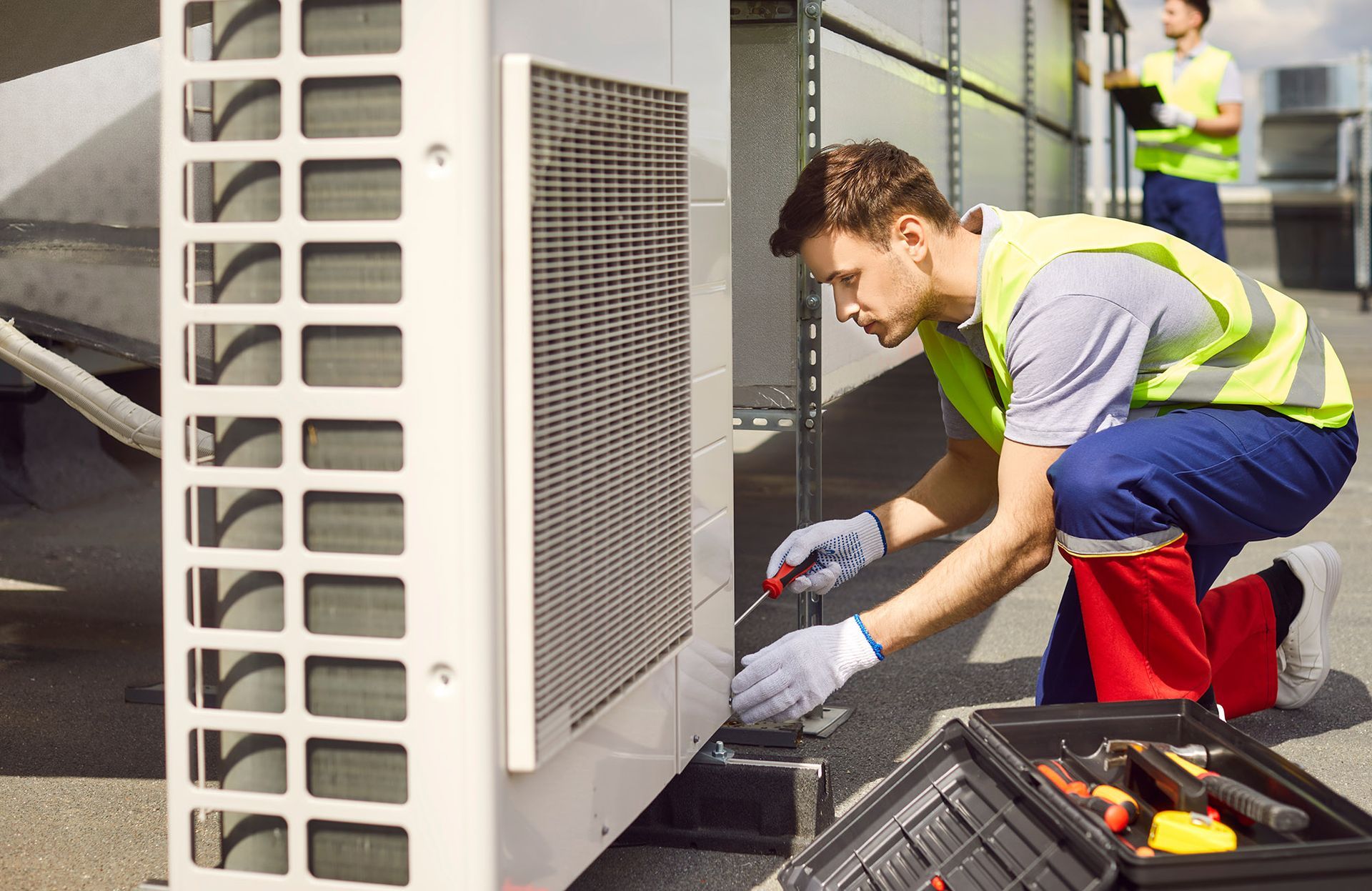 Two HVAC technicians working on rooftop air conditioning unit, one kneeling, using a screwdriver.