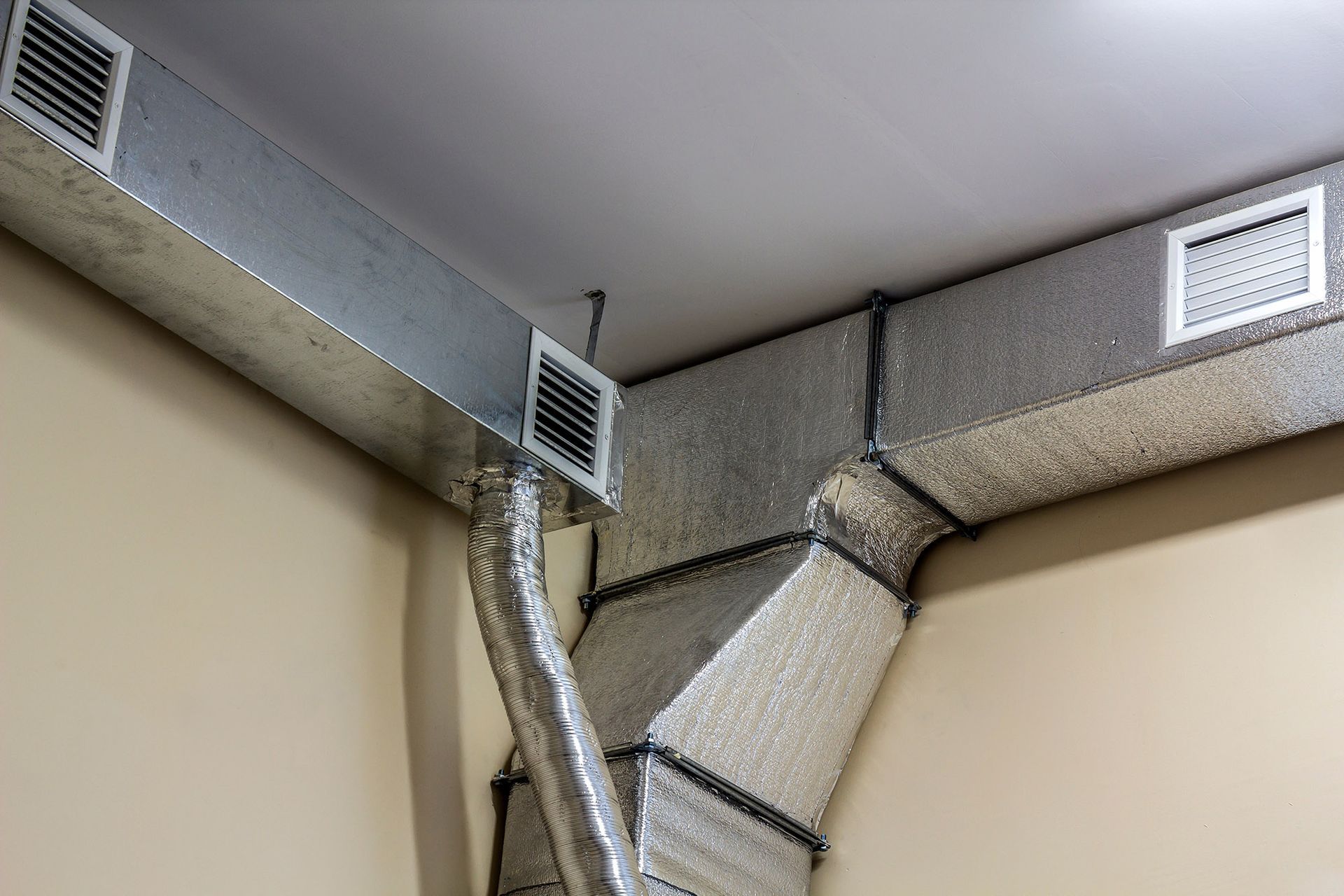 HVAC ductwork installation in a building corner, with silver metal ducts and vents against beige walls and white ceiling.