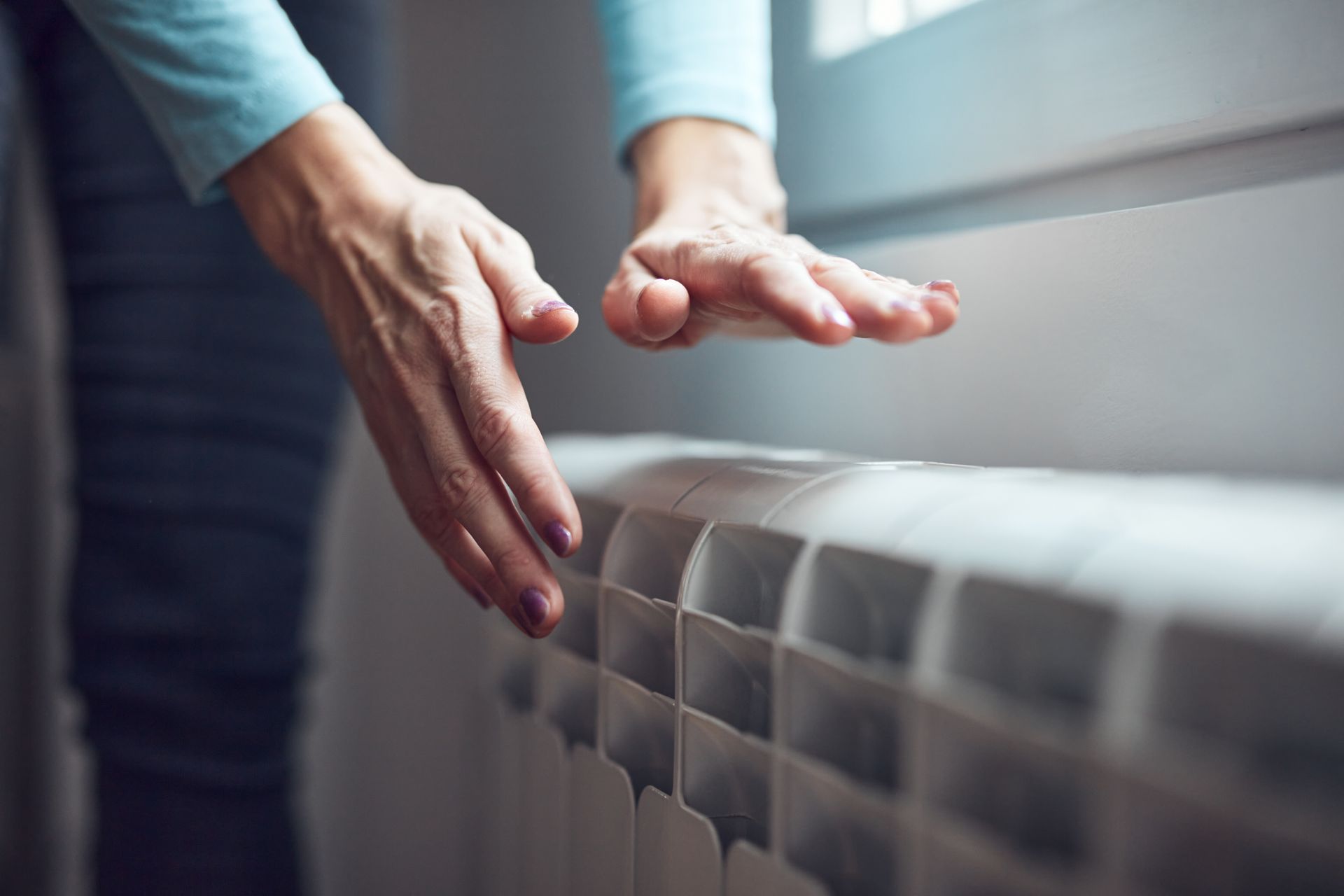 Hands hovering over a white radiator near a window, suggesting heat adjustment or checking warmth Hands hovering over a white radiator near a window, suggesting heat adjustment or checking warmth