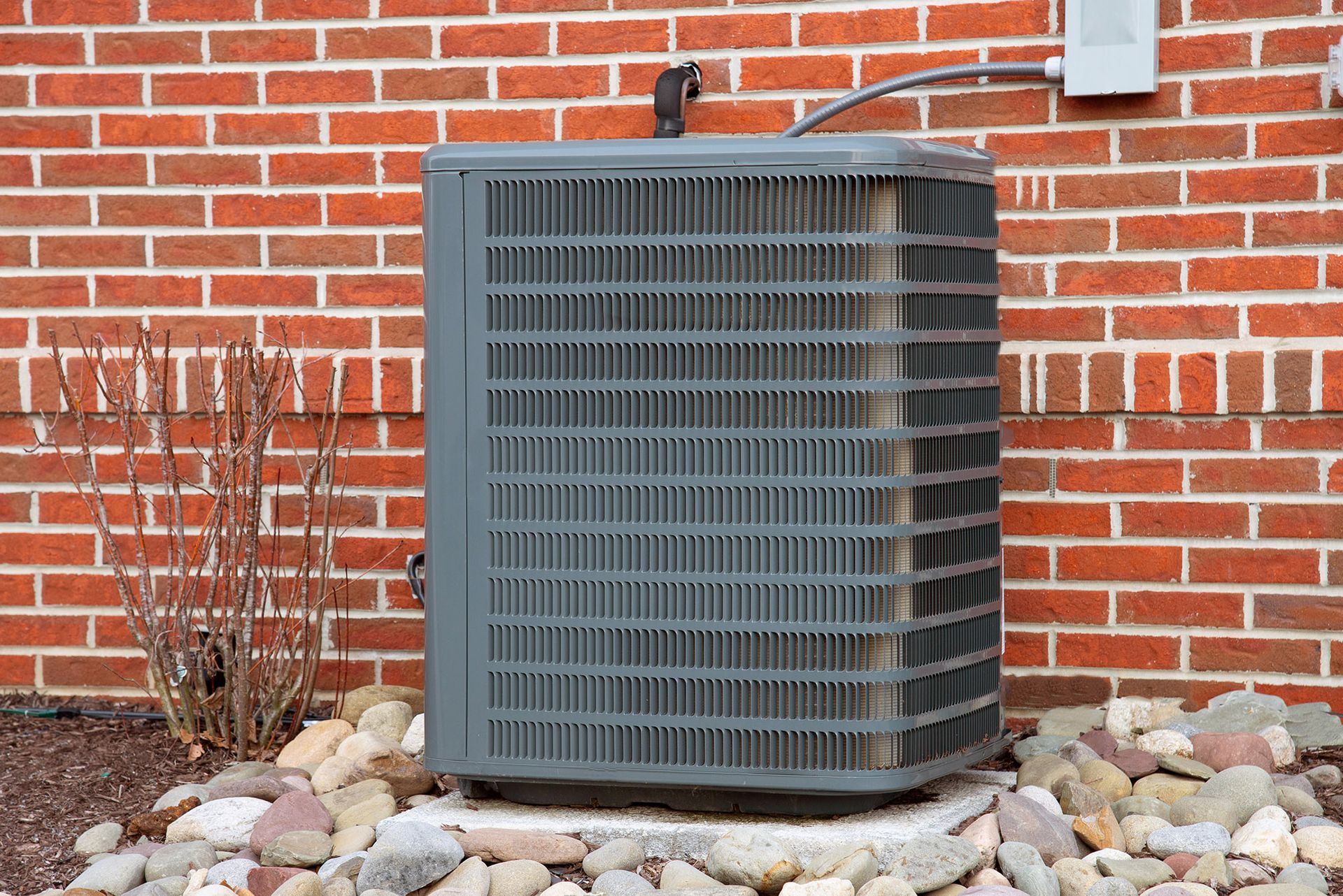 An outdoor air conditioning unit against a red brick wall, surrounded by rocks and a small bush.