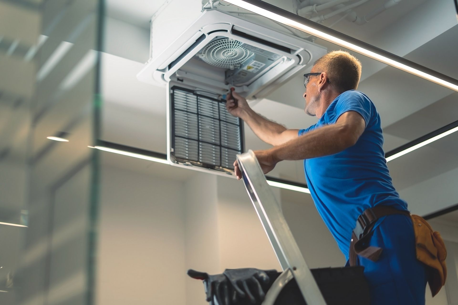 Man on ladder cleaning an air conditioning unit filter indoors.