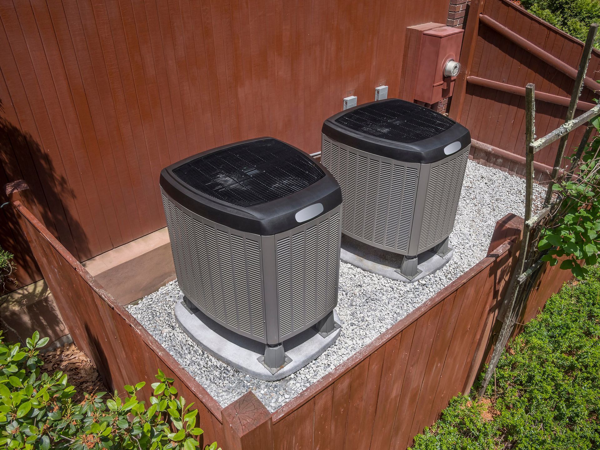 Two air conditioning units on a gravel base next to a wood structure.