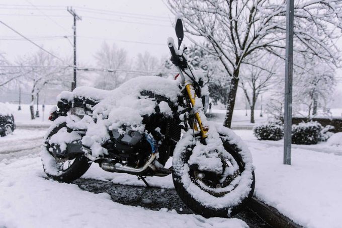 A motorcycle covered in a thick layer of snow, parked on a roadside during a winter storm with bare trees in the background.