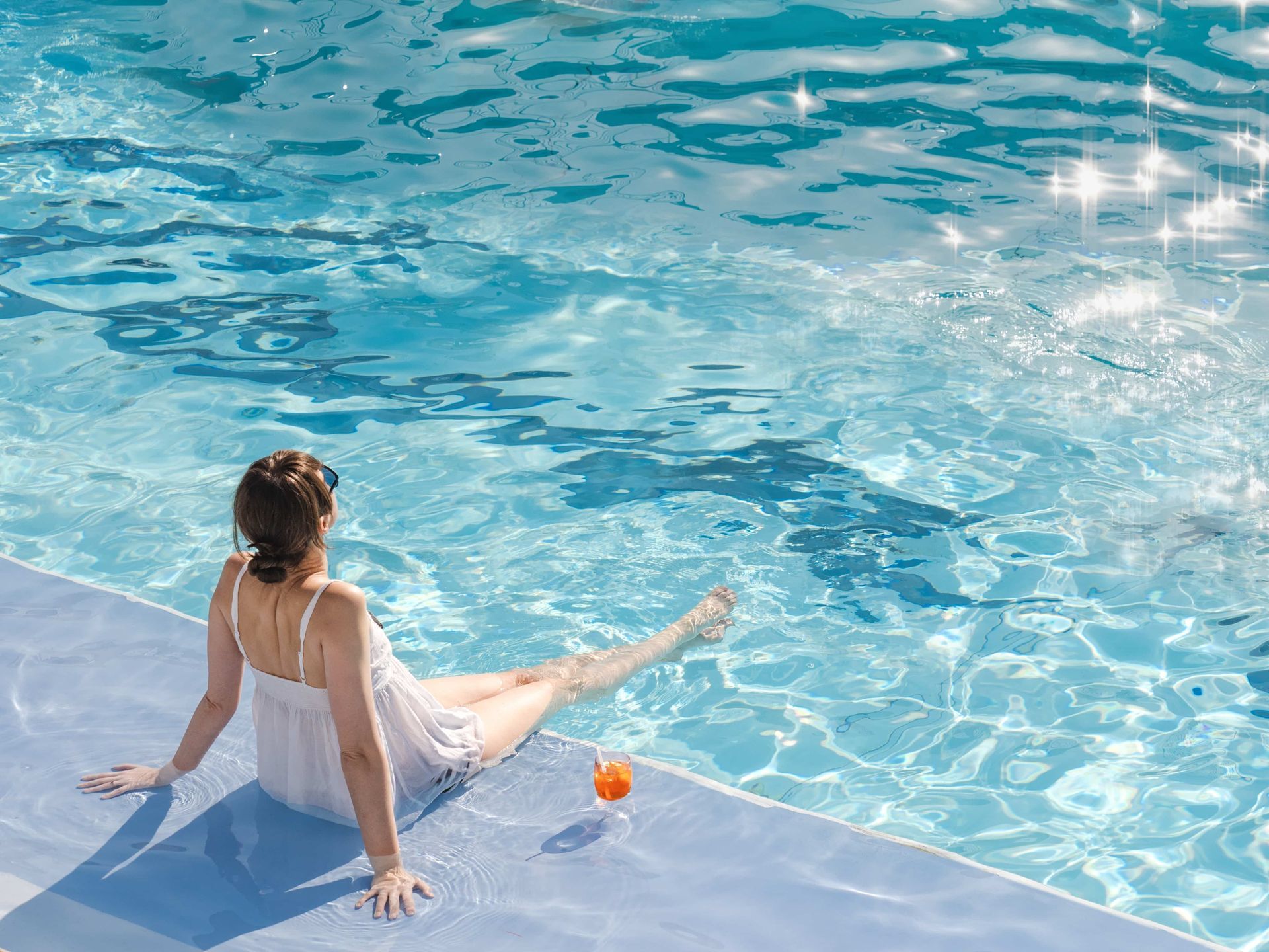 Woman relaxing by pool, legs in water, drink beside her.