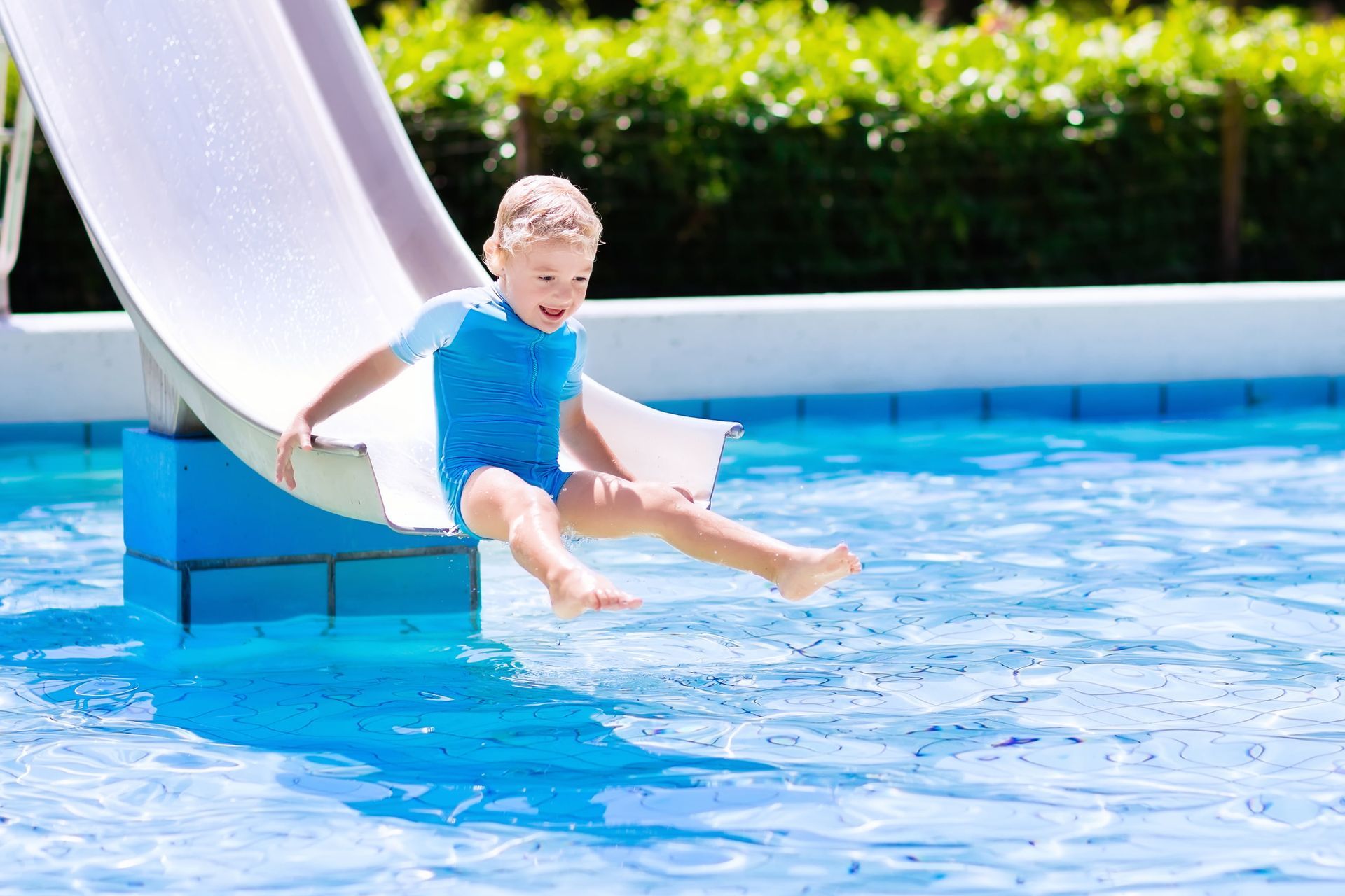 Boy in blue swimsuit sliding down a pool slide into water.