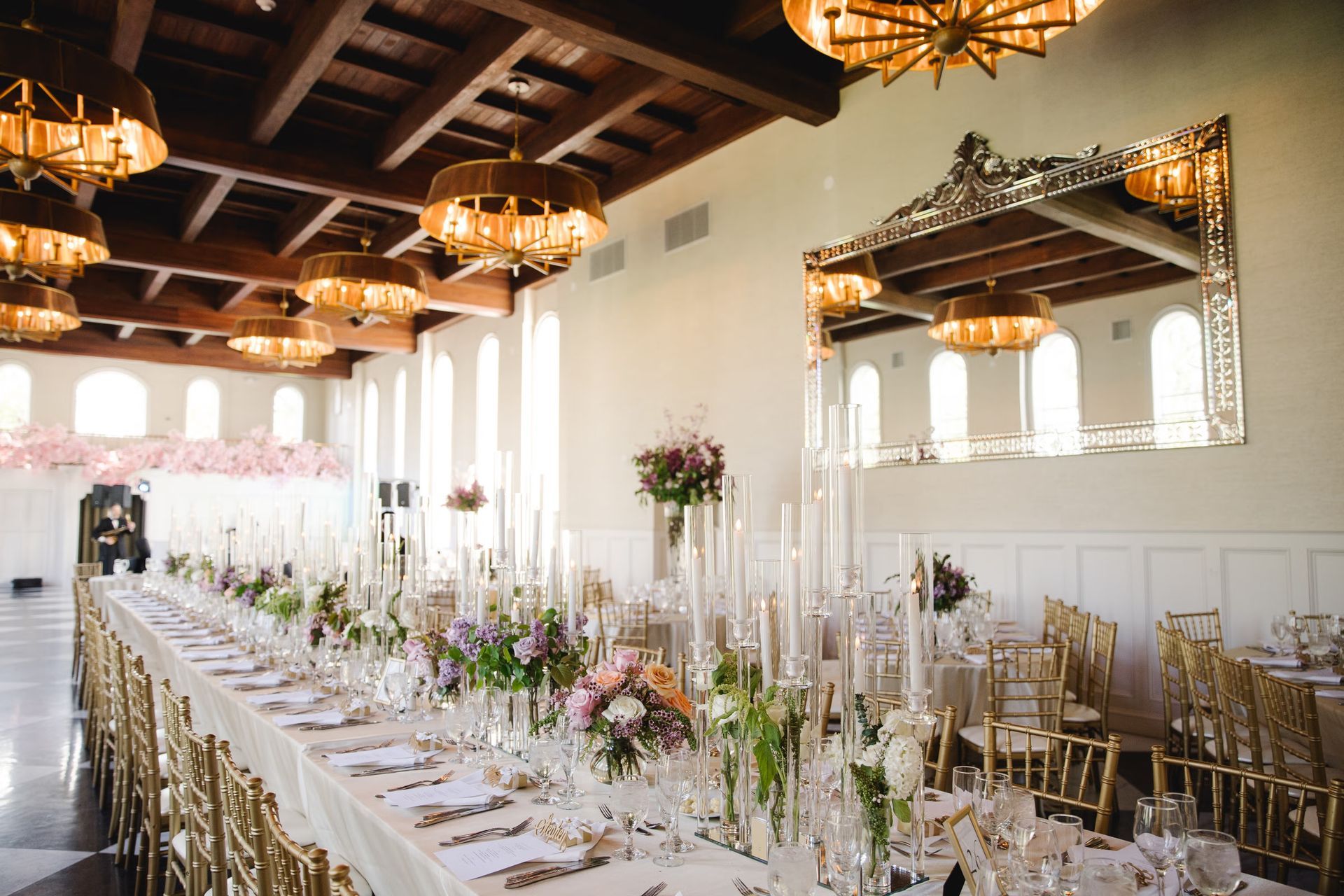 Elegant wedding reception hall with long banquet tables, gold chairs, chandeliers, and floral centerpieces