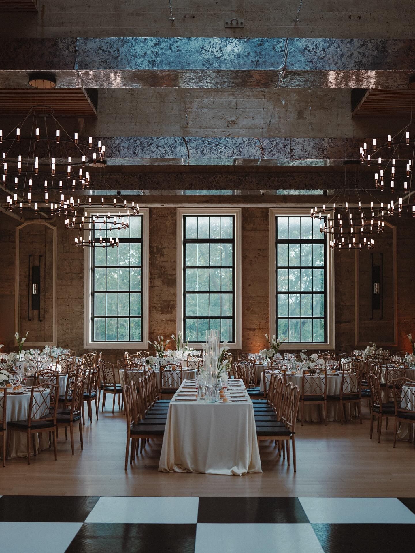Elegant banquet hall with chandeliers, long tables, and a white-draped table centered by tall windows.