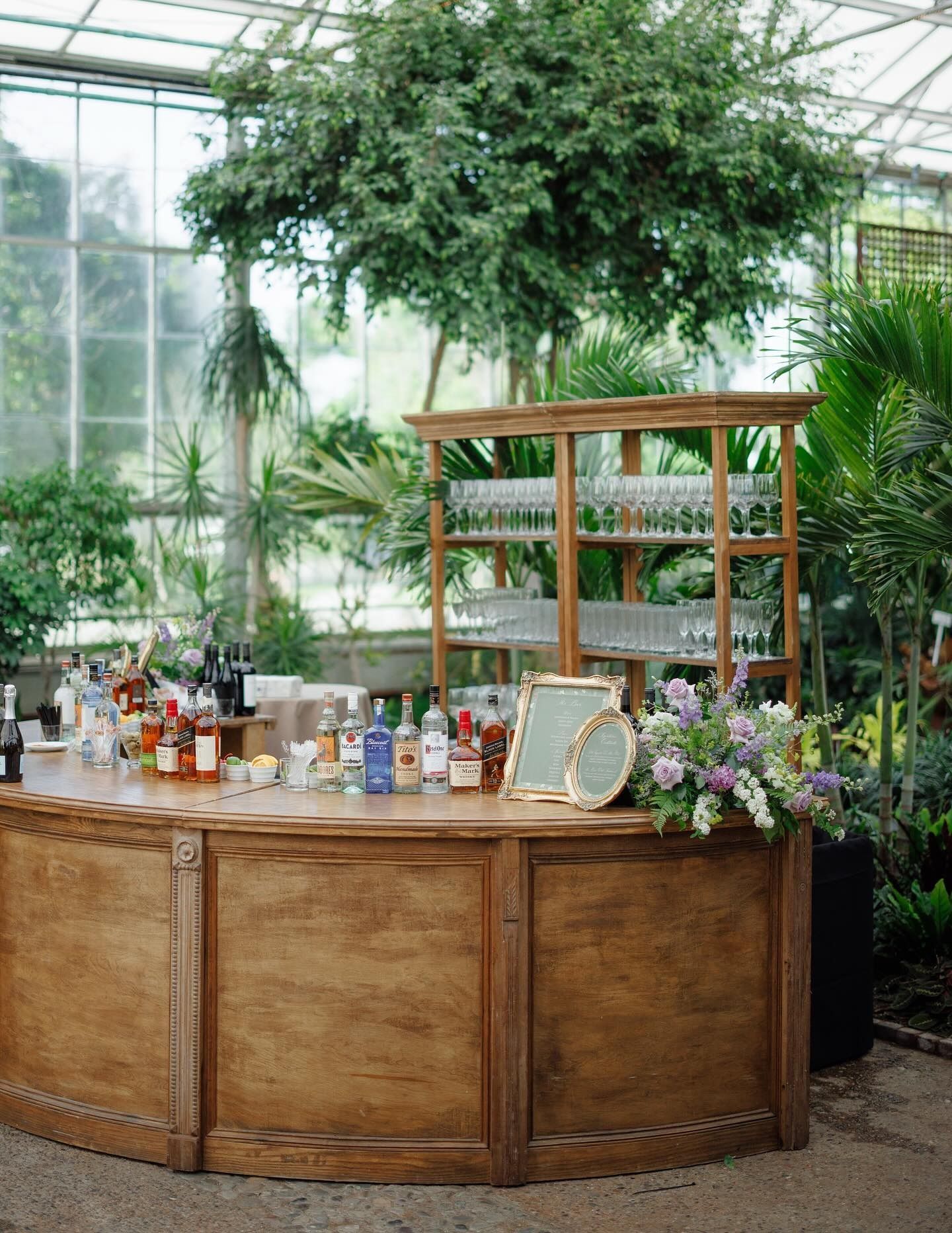 Wooden bar in a lush greenhouse with plants, flowers, and bottles on the counter