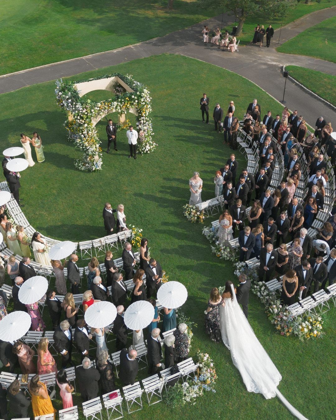 Outdoor wedding ceremony with bride, groom, guests, and musicians on a green lawn under a floral arch