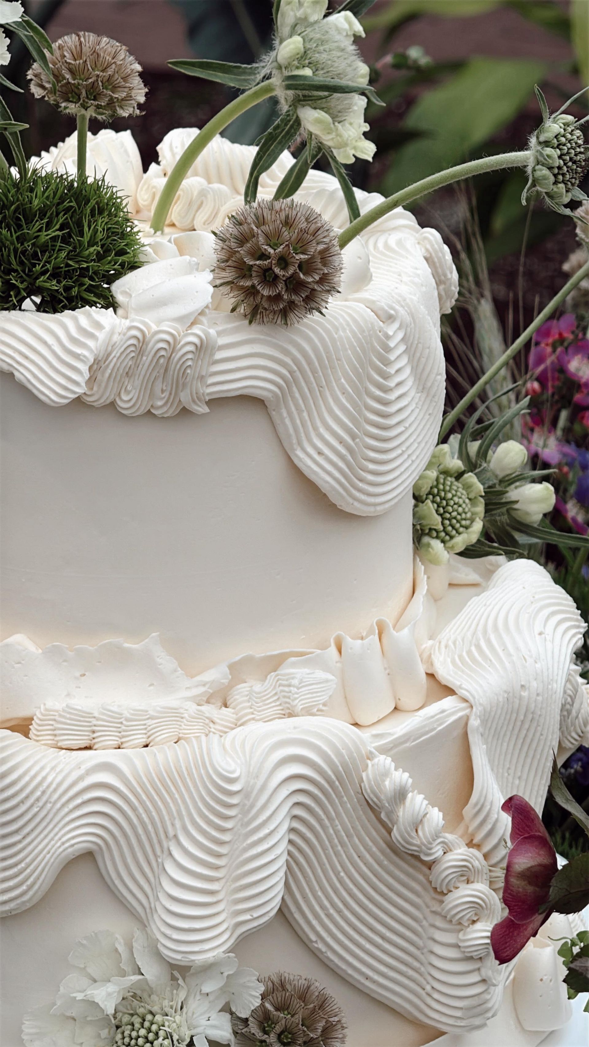 White frosted tiered cake with wavy piping and dried flowers against a dark floral background