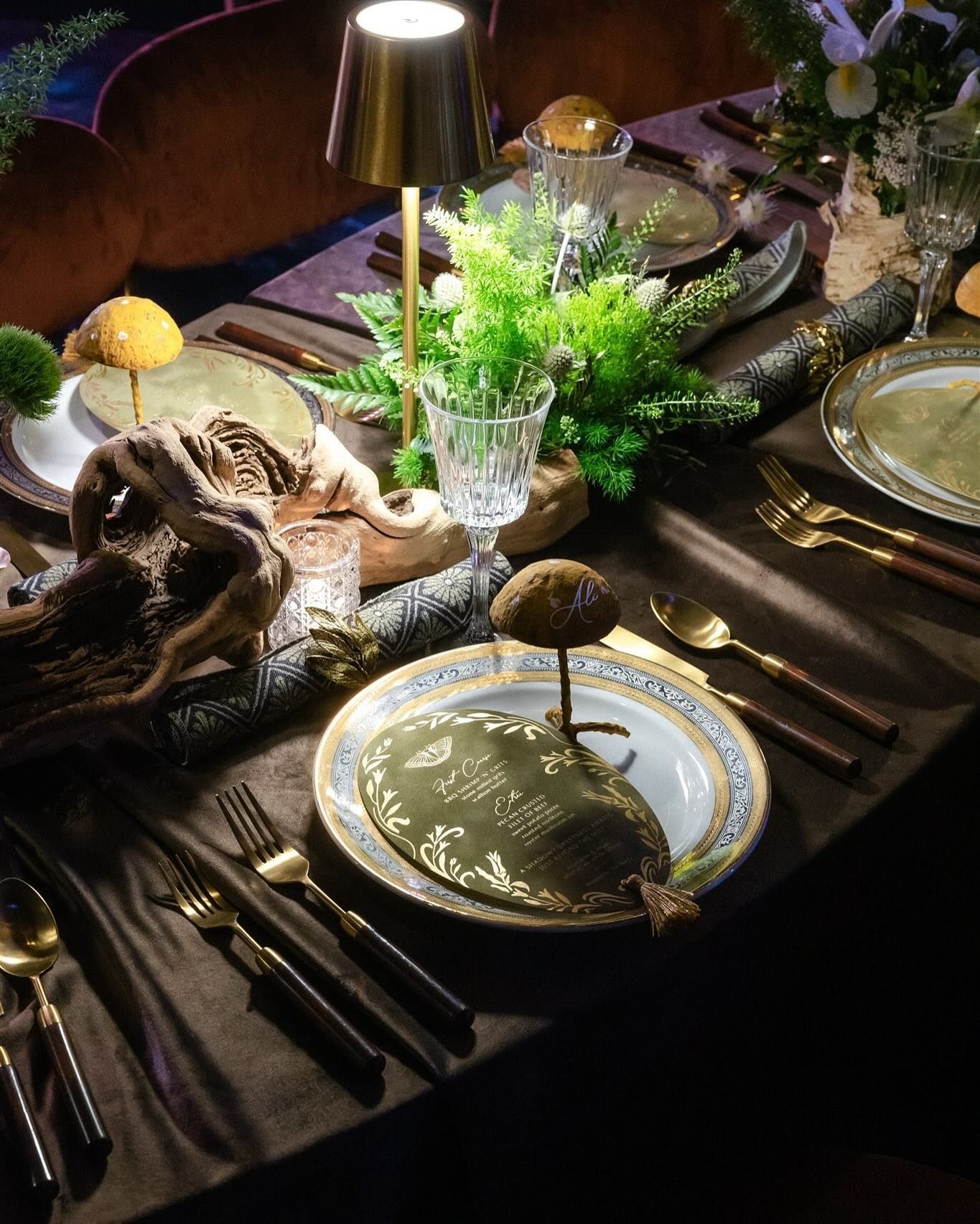 Elegant dining table set with gold-rimmed plates, candles, greenery, and dark tableware in warm light